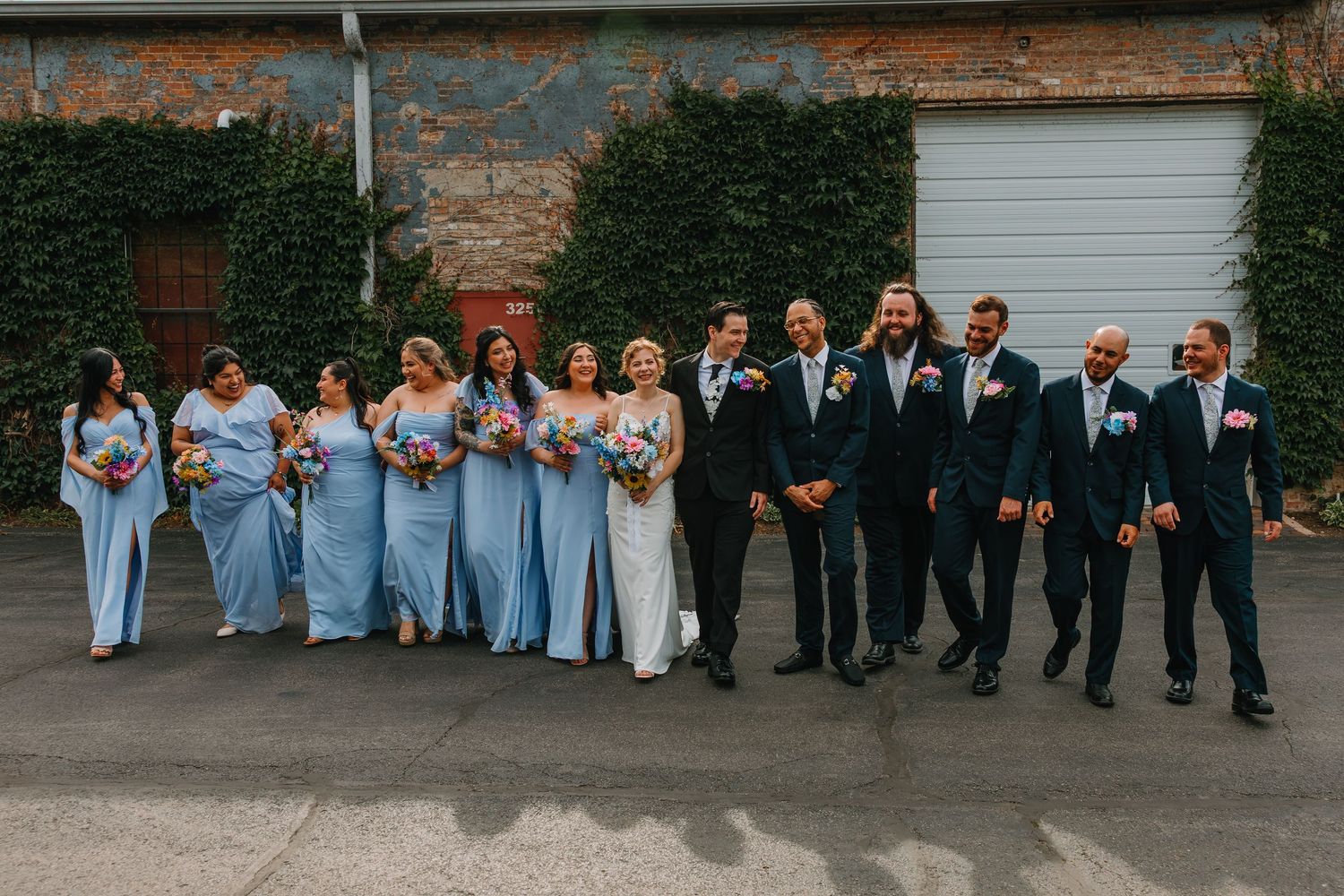 Full wedding party in light blue bridesmaid gowns and navy suits poses casually outside brick venue. Chicagoland wedding venues.