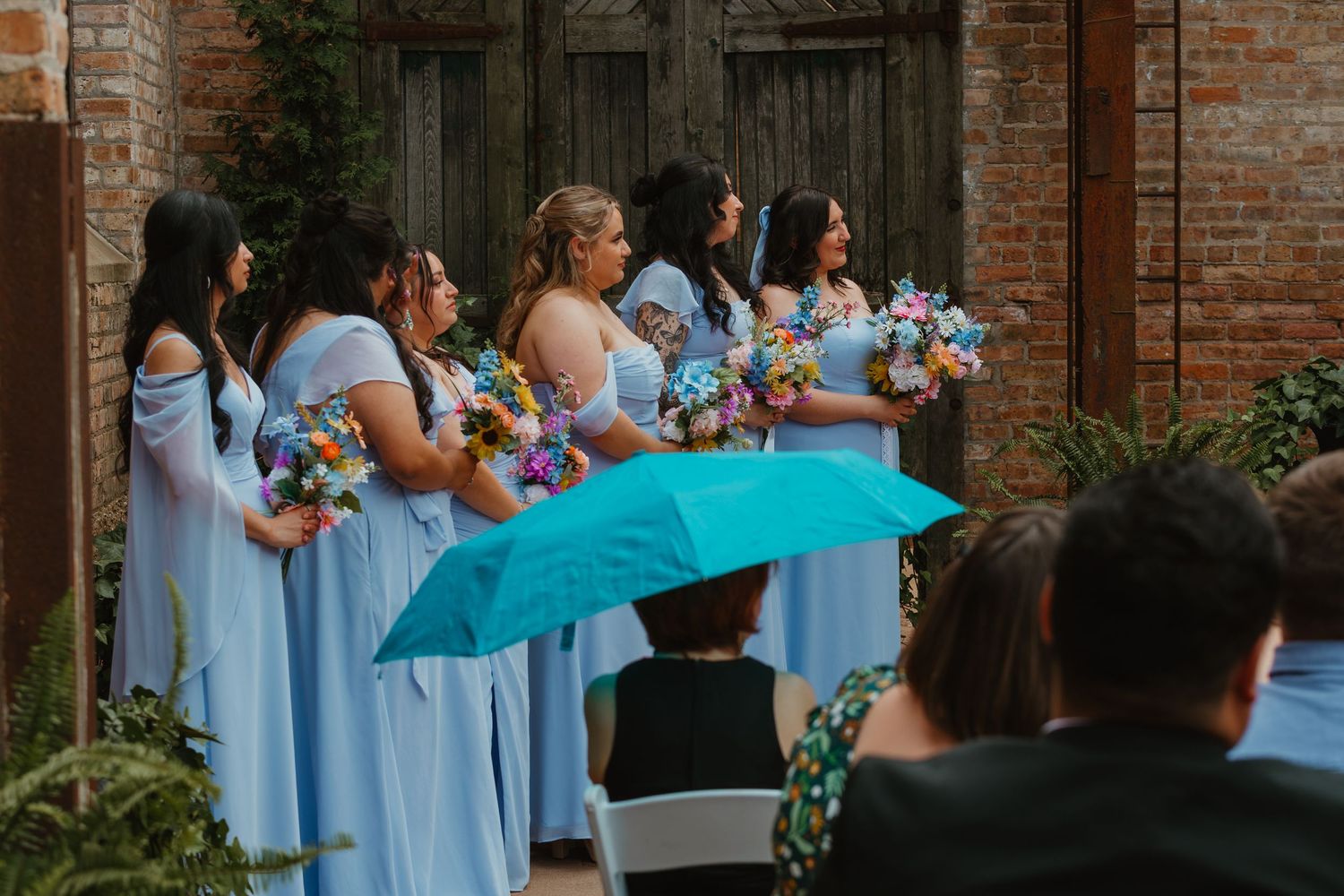 Bridesmaids in light blue gowns holding colorful bouquets stand in line as guest uses teal umbrella at ceremony. Chicagoland wedding venues.