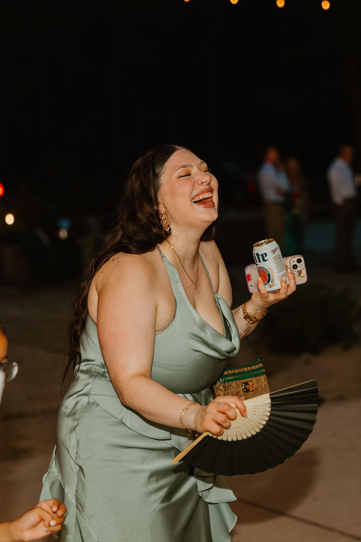 Joyful woman in sage green satin dress laughing while holding a beer can and decorative fan at night. Northwest Indiana wedding venues.