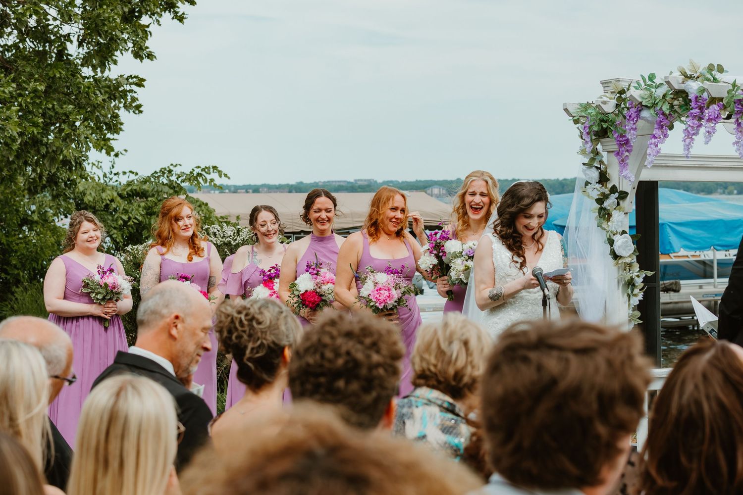 Bridesmaids in purple dresses smile joyfully beside bride during outdoor lakeside wedding ceremony under floral arch.