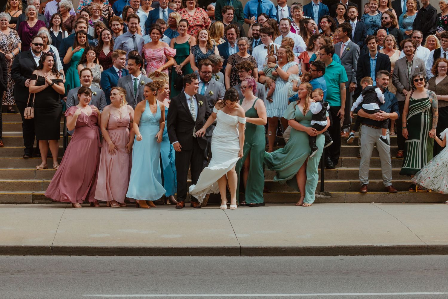 Large wedding party group photo on outdoor steps with bridesmaids in pink and teal gowns.