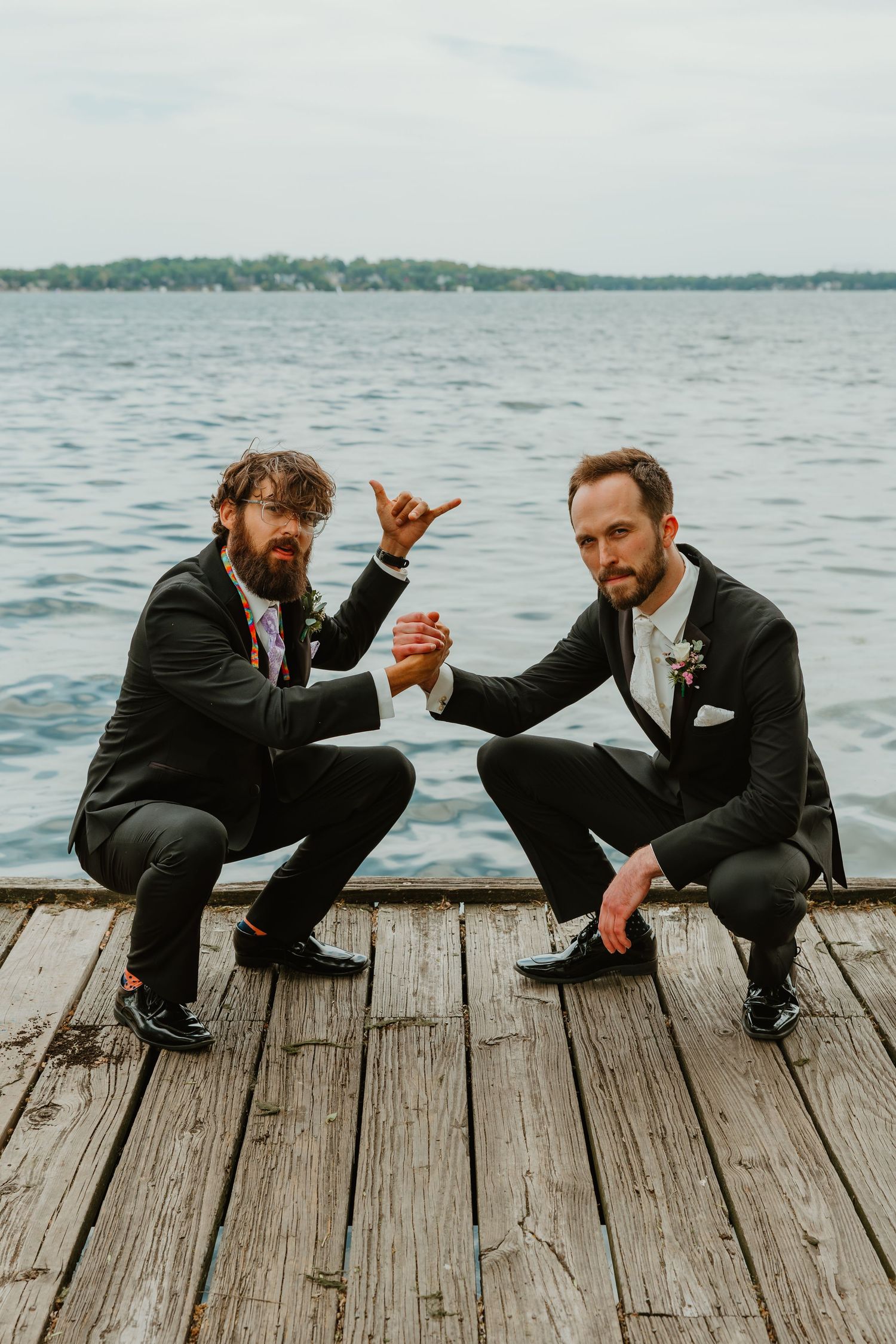 Two groomsmen in black suits crouching on a wooden dock giving a fun pose by the lake.