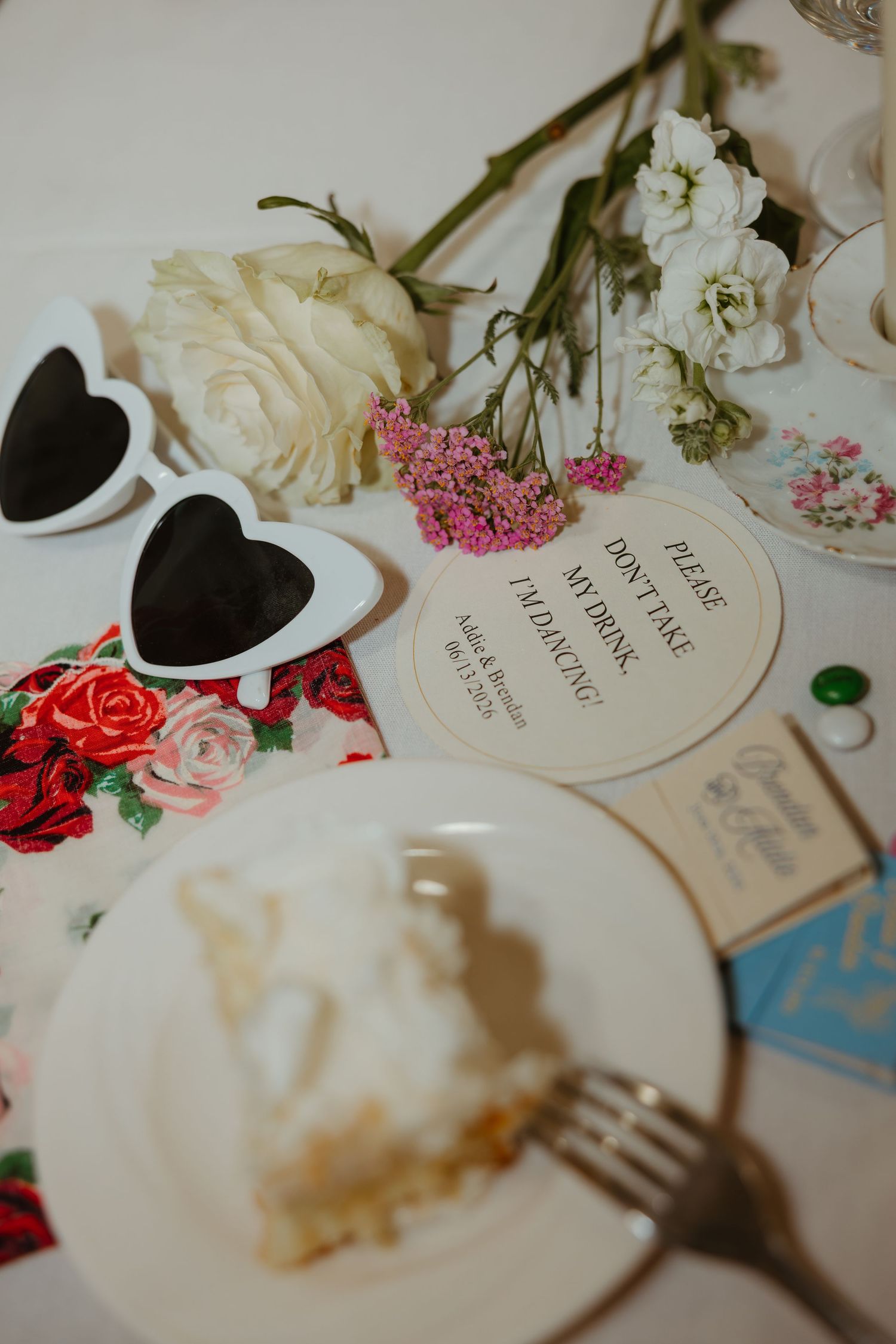 Wedding reception table with heart sunglasses, white roses, cake slice, and drink marker sign. Detailed late night wedding flat lay.