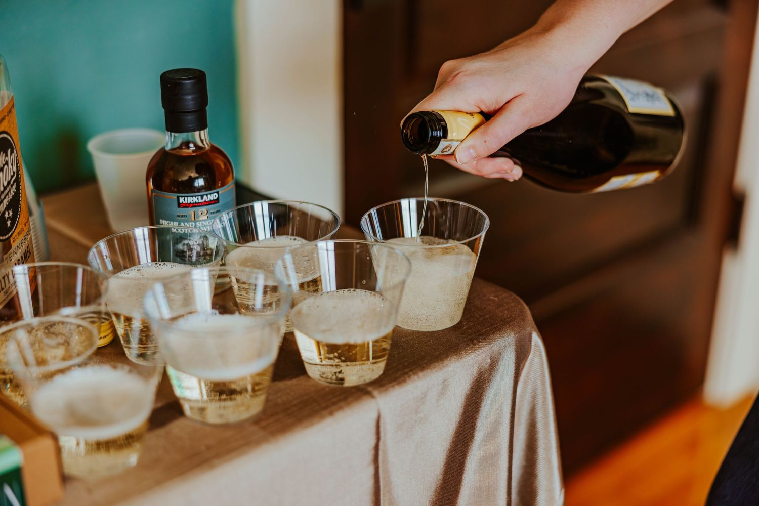 Hand pouring champagne into clear plastic cups on linen-covered table at wedding reception.