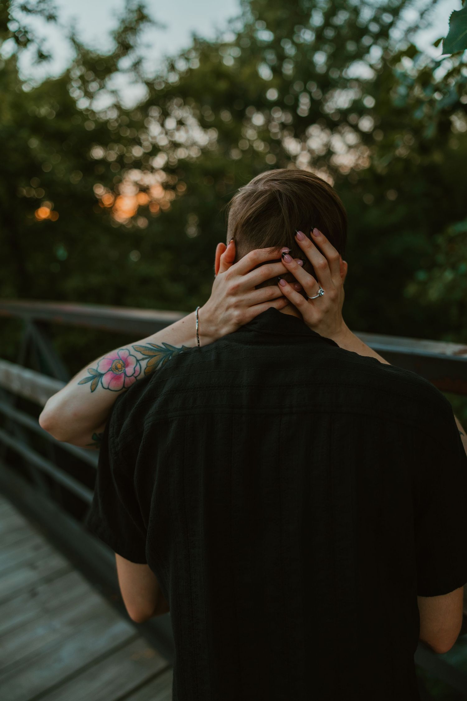 Tattooed woman's hand with engagement ring resting on man's neck from behind on a wooden bridge at sunset. Chicagoland engagement photos.