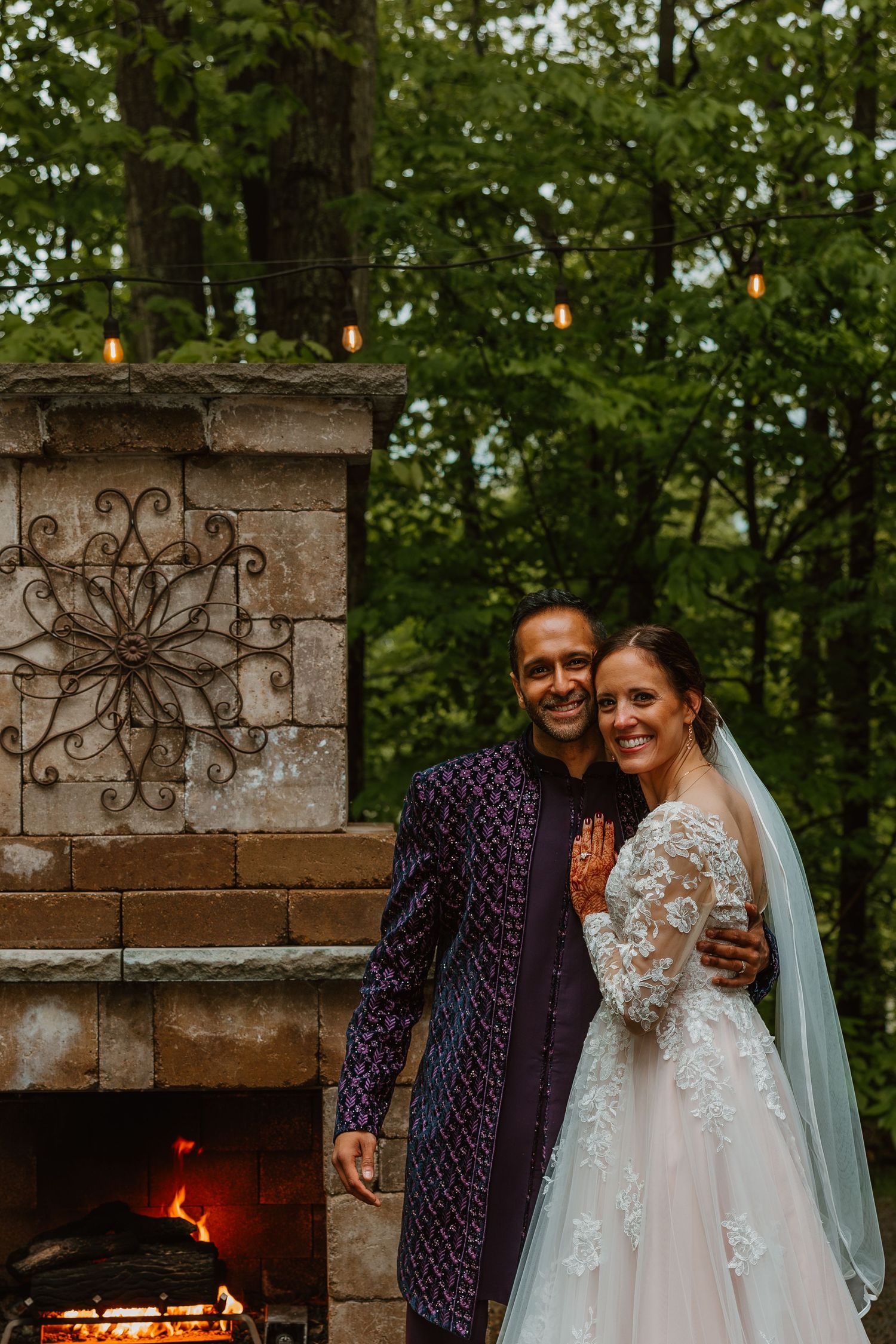 Bride and groom smiling together by a stone outdoor fireplace surrounded by lush green trees. Documentary style wedding photos.