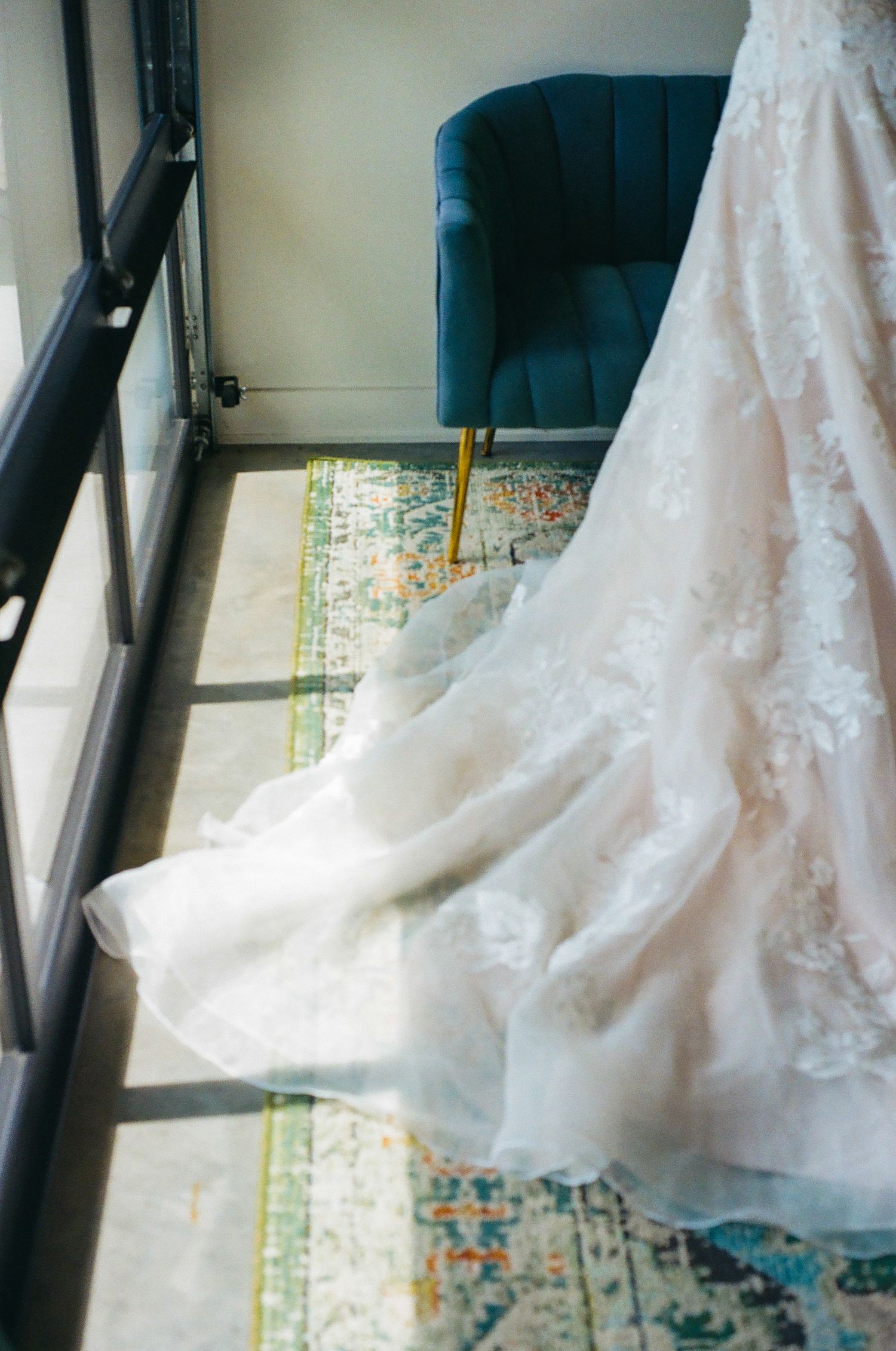 Elegant white lace wedding dress train pooling on floor near window with blue velvet chair in background.