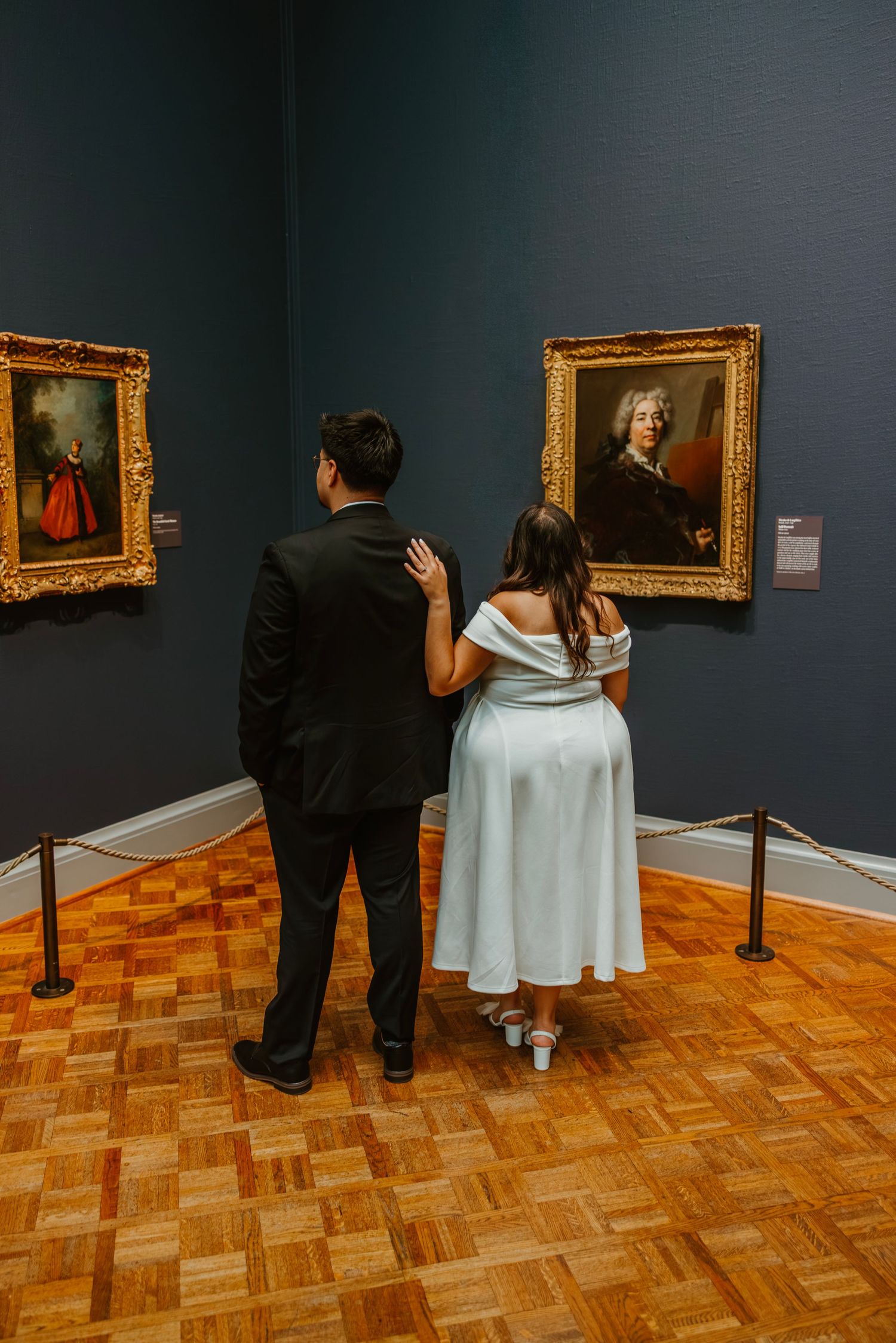 Couple in wedding attire viewing classic portrait paintings in elegant museum gallery with wood floors. Engagement at Chicago Art Institute.