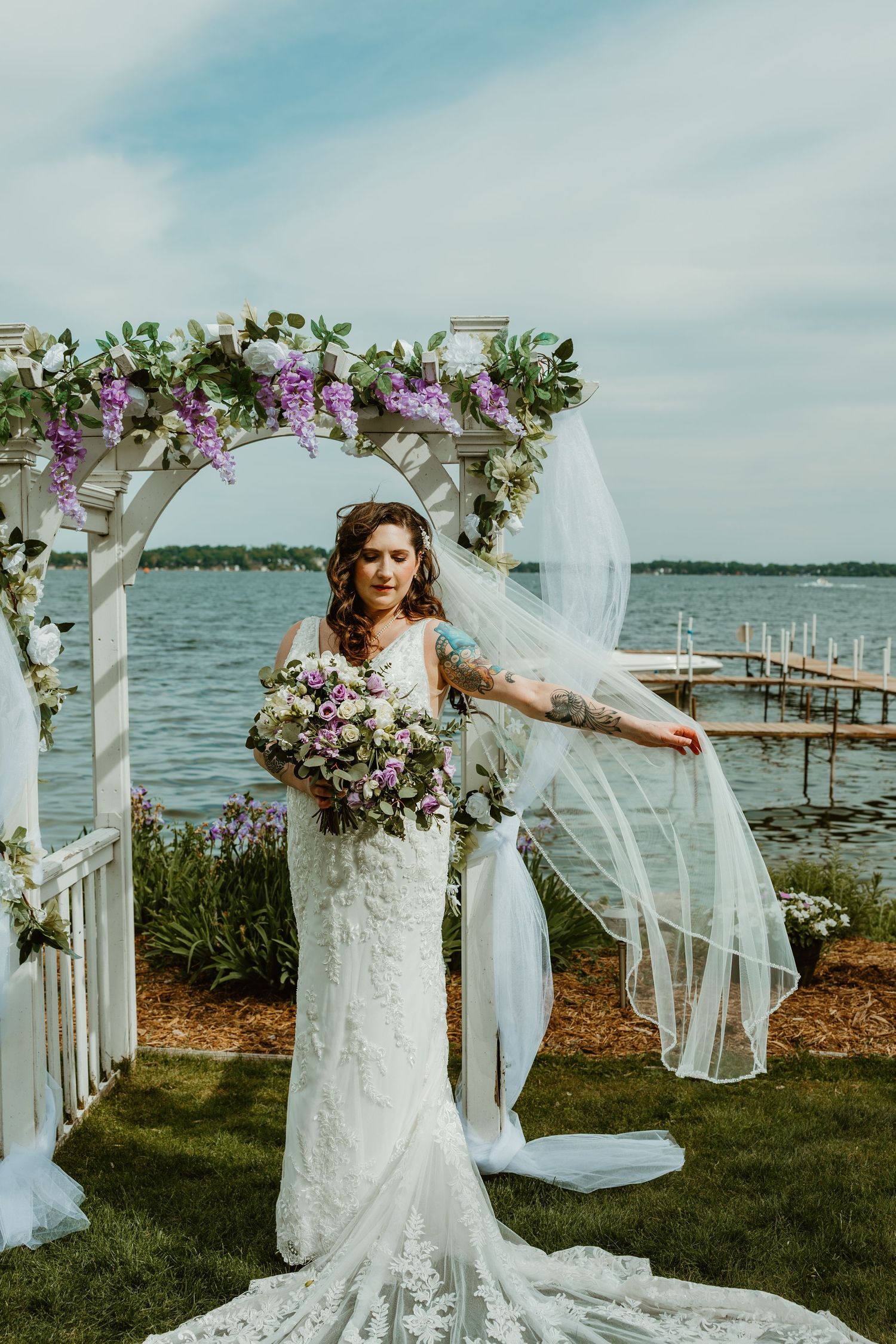 Bride in lace gown with flowing veil poses under white floral arch beside serene lake and dock.