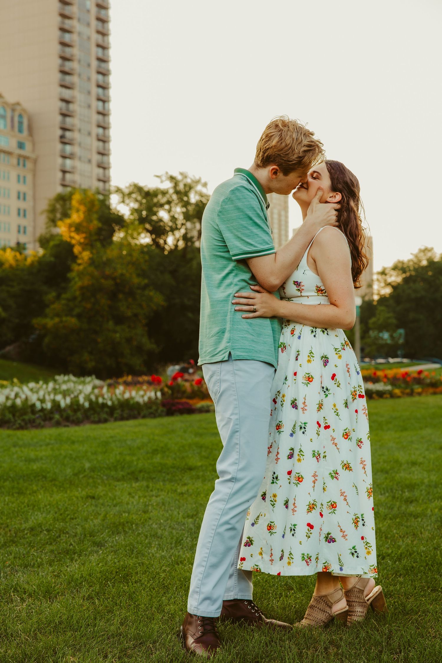 Couple sharing a romantic kiss on a green lawn with flower gardens and a city building in the background. Chicago engagement photos.