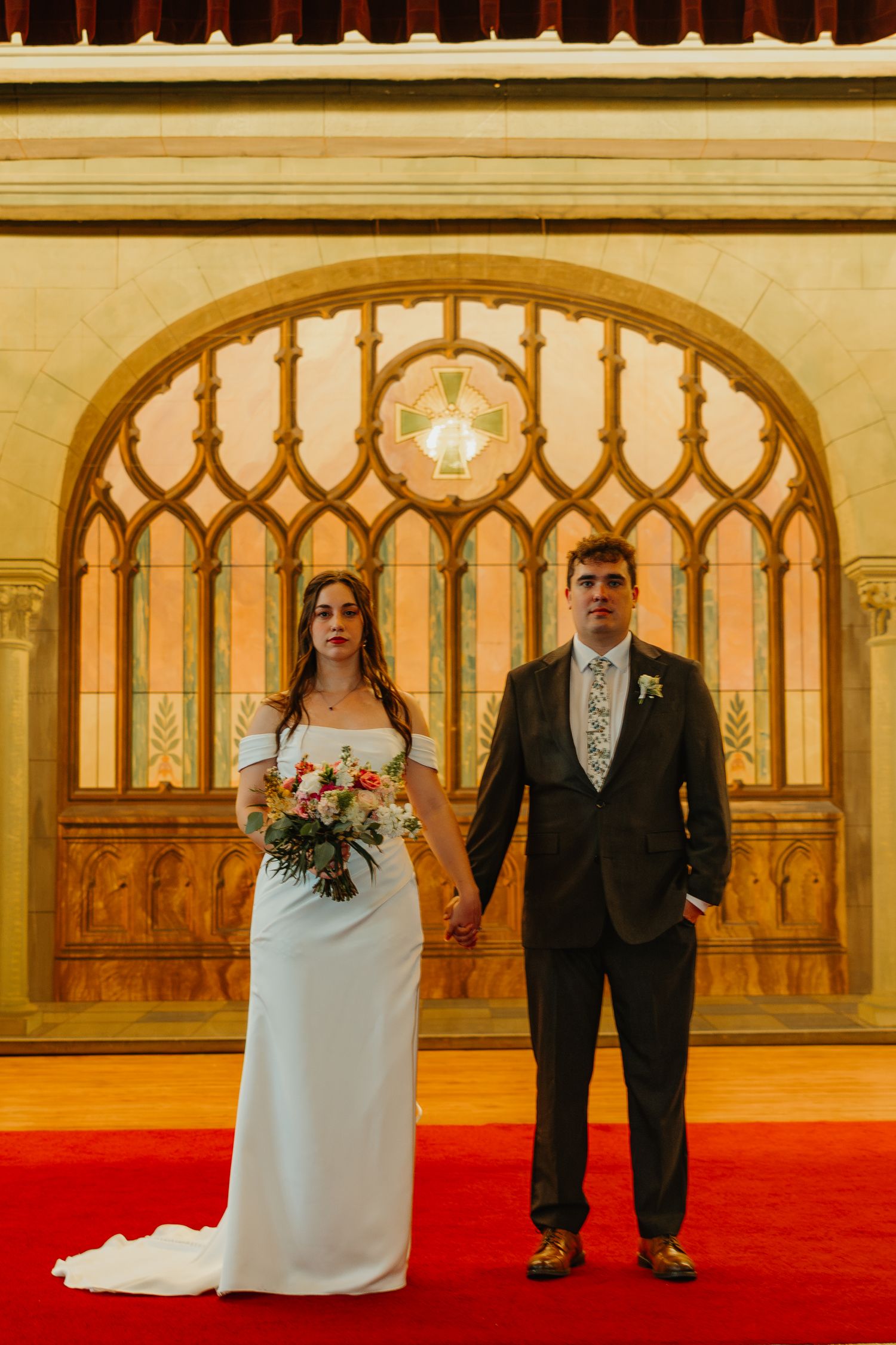 Bride and groom holding hands standing before ornate stained glass arch on red carpet in church.