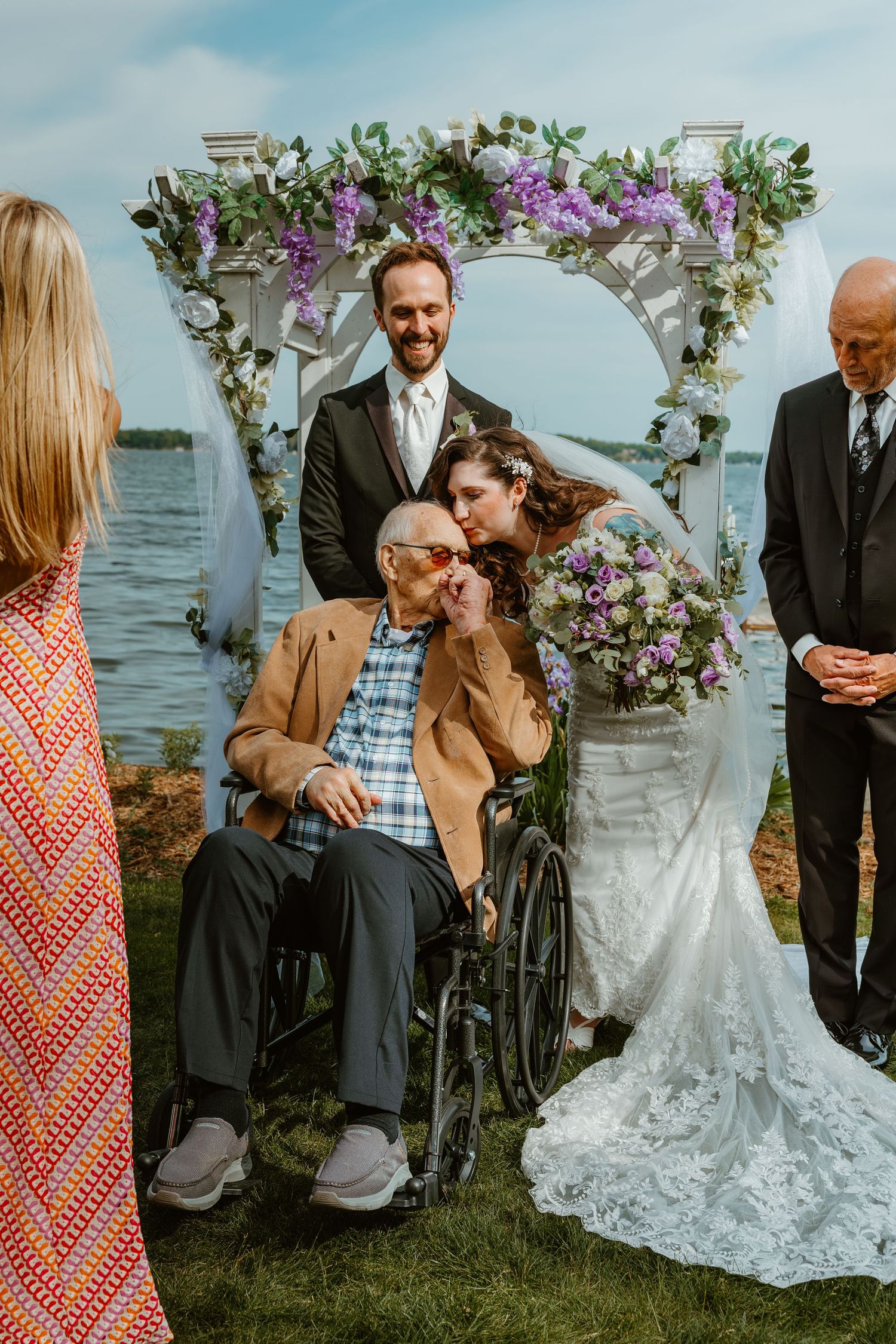 Bride in lace gown kisses elderly man in wheelchair under a floral arch at lakeside wedding.
