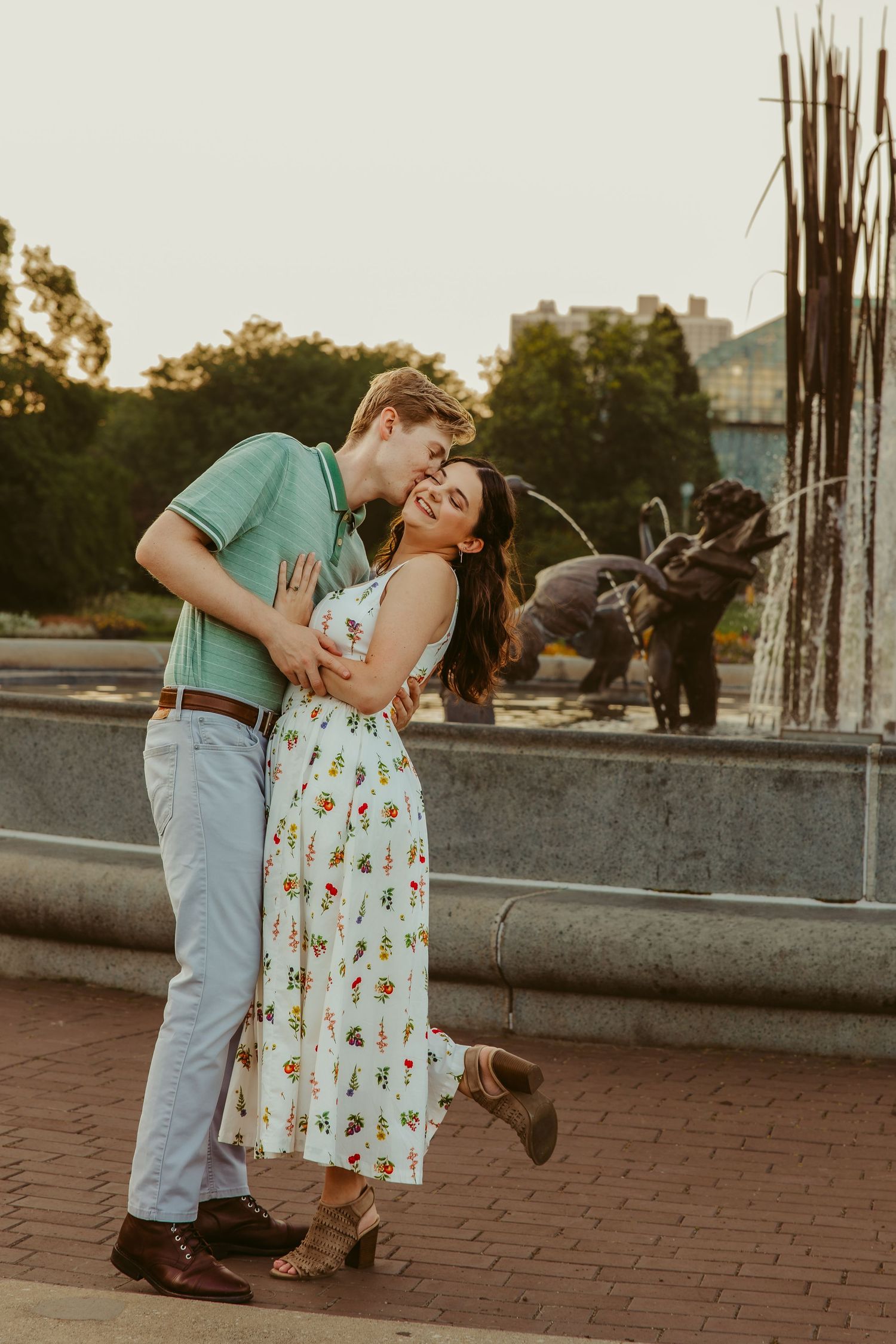Couple laughing and embracing near a fountain at golden hour, woman lifting her foot joyfully. Chicago engagement photos.