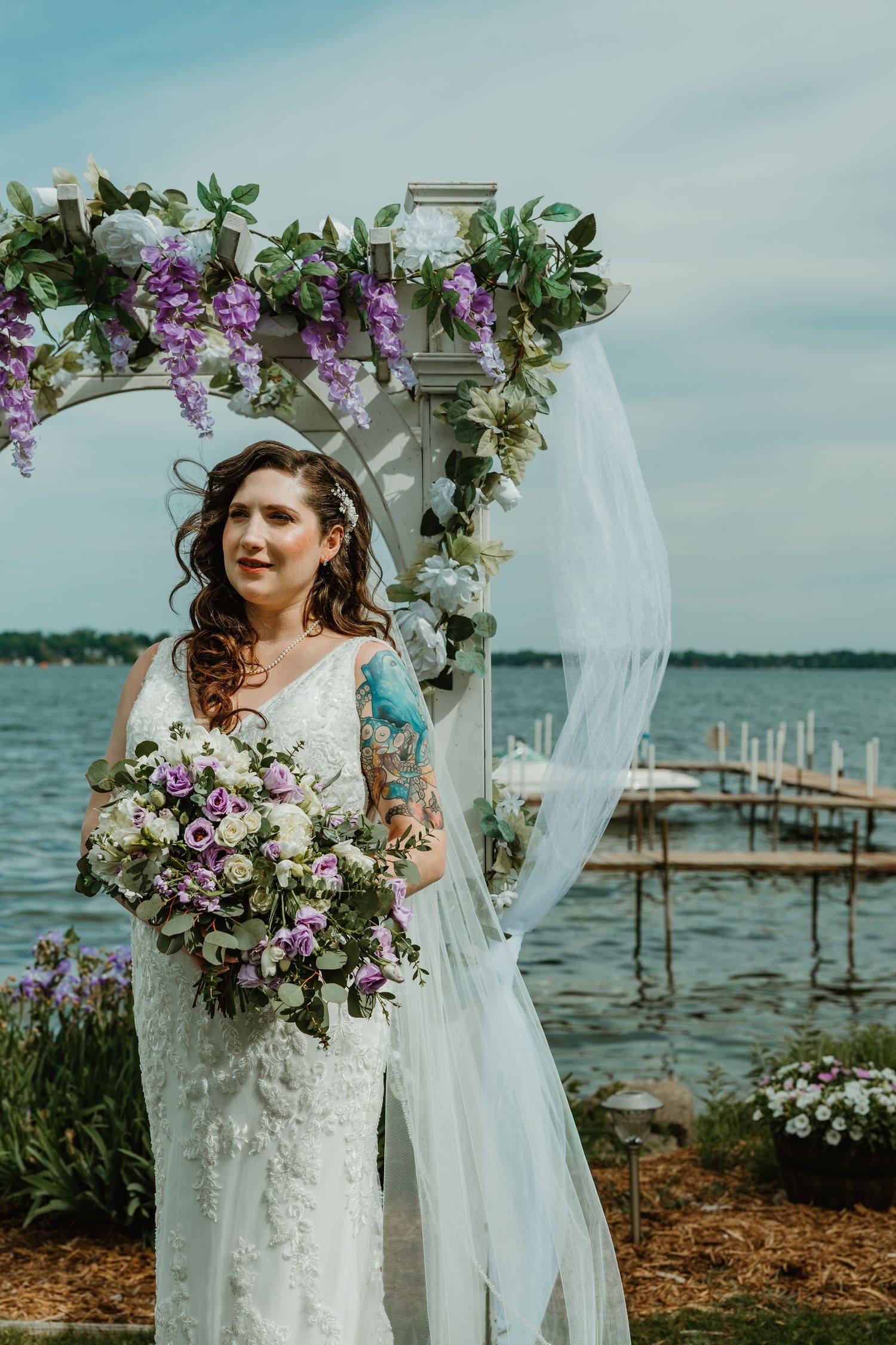 Bride in lace gown with flowing veil smiles holding purple bouquet under wisteria arch at lakeside wedding.