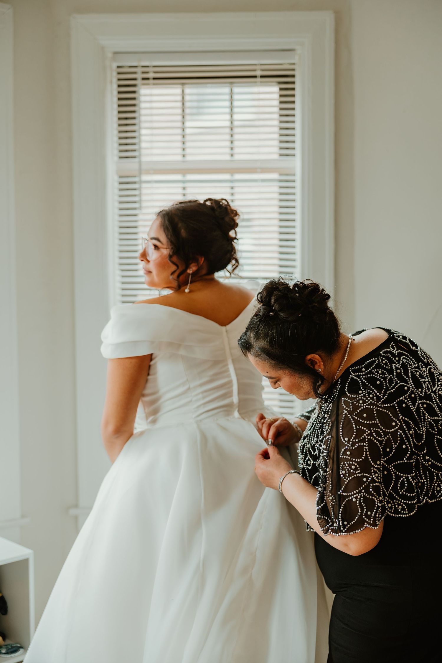 Woman in beaded black dress buttons the back of bride's off-shoulder white wedding gown by window. Chicagoland weddings.
