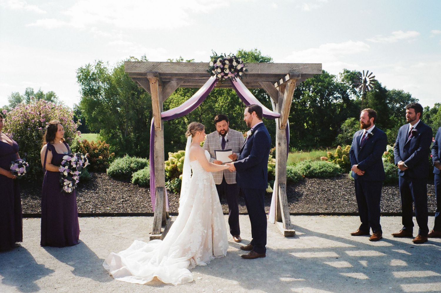 Outdoor wedding ceremony under a wooden arch with purple floral decor, bride and groom exchanging vows with wedding party.