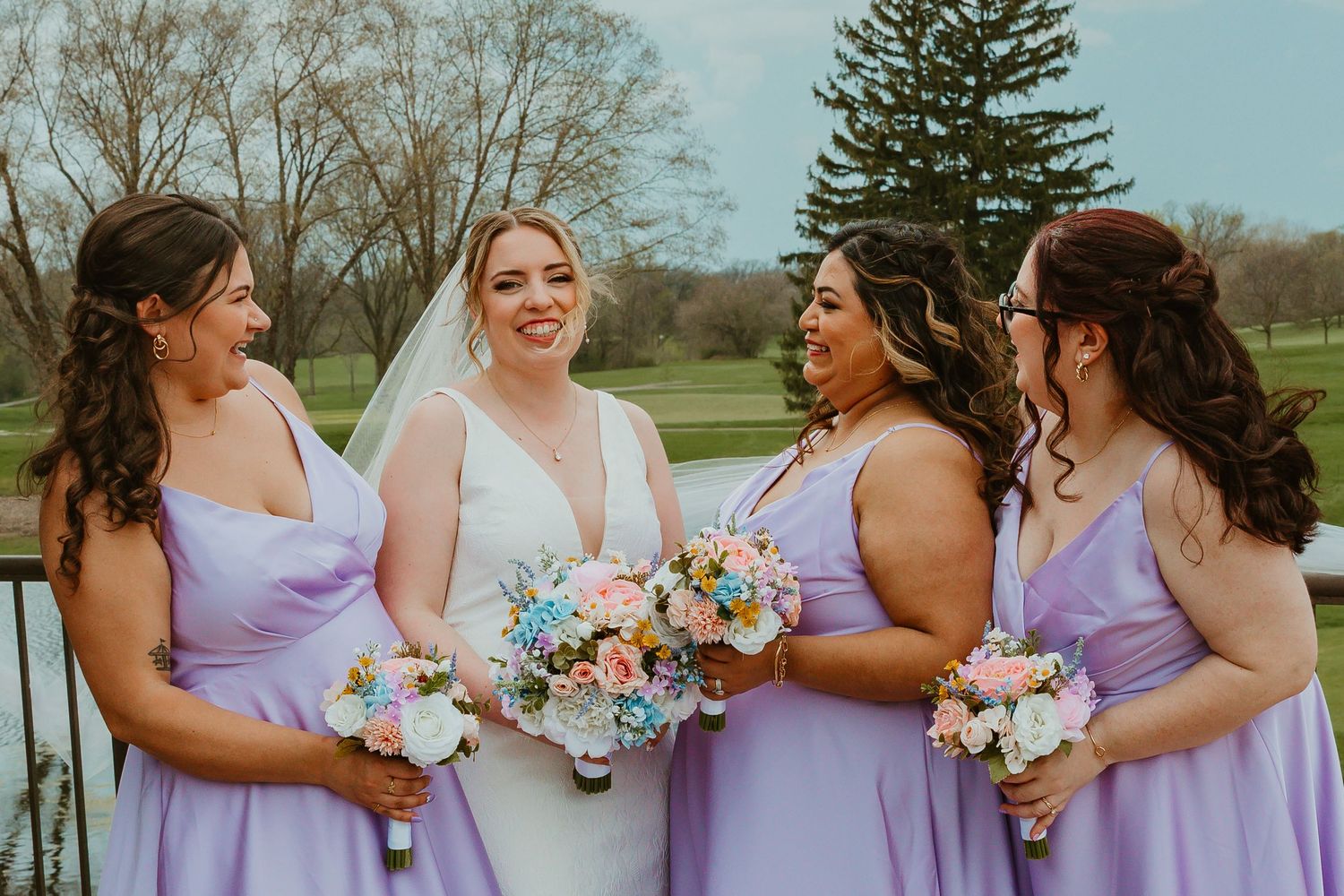 Bride laughing with three bridesmaids in lavender dresses holding colorful bouquets outdoors. Chicagoland wedding venues.