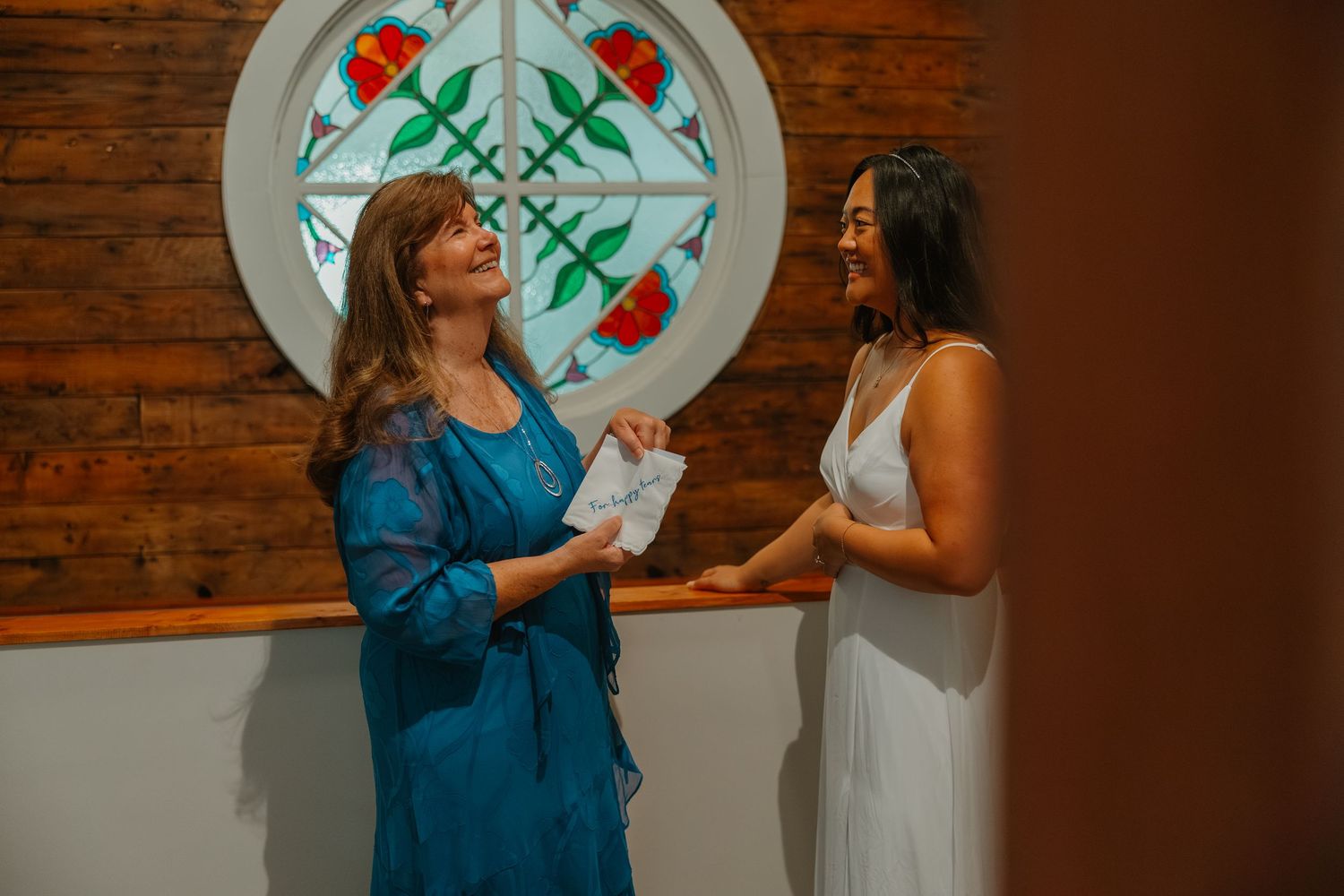 Two women in a church, one in blue and one in white, sharing a joyful moment by a stained glass window.