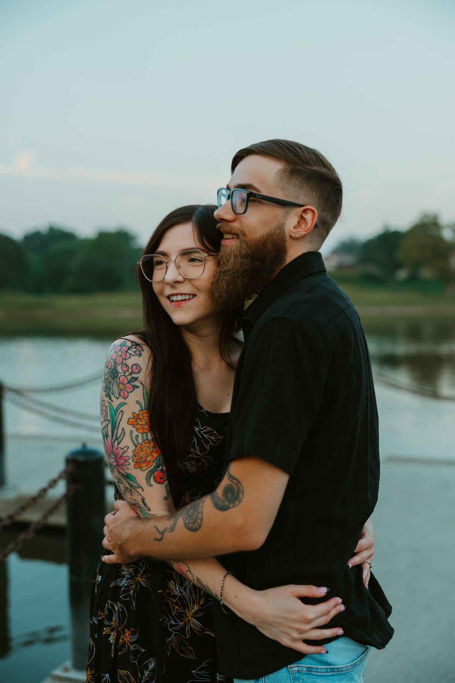 Tattooed couple embracing near a lakeside dock at dusk, both wearing glasses, smiling joyfully. Chicagoland engagement photos.