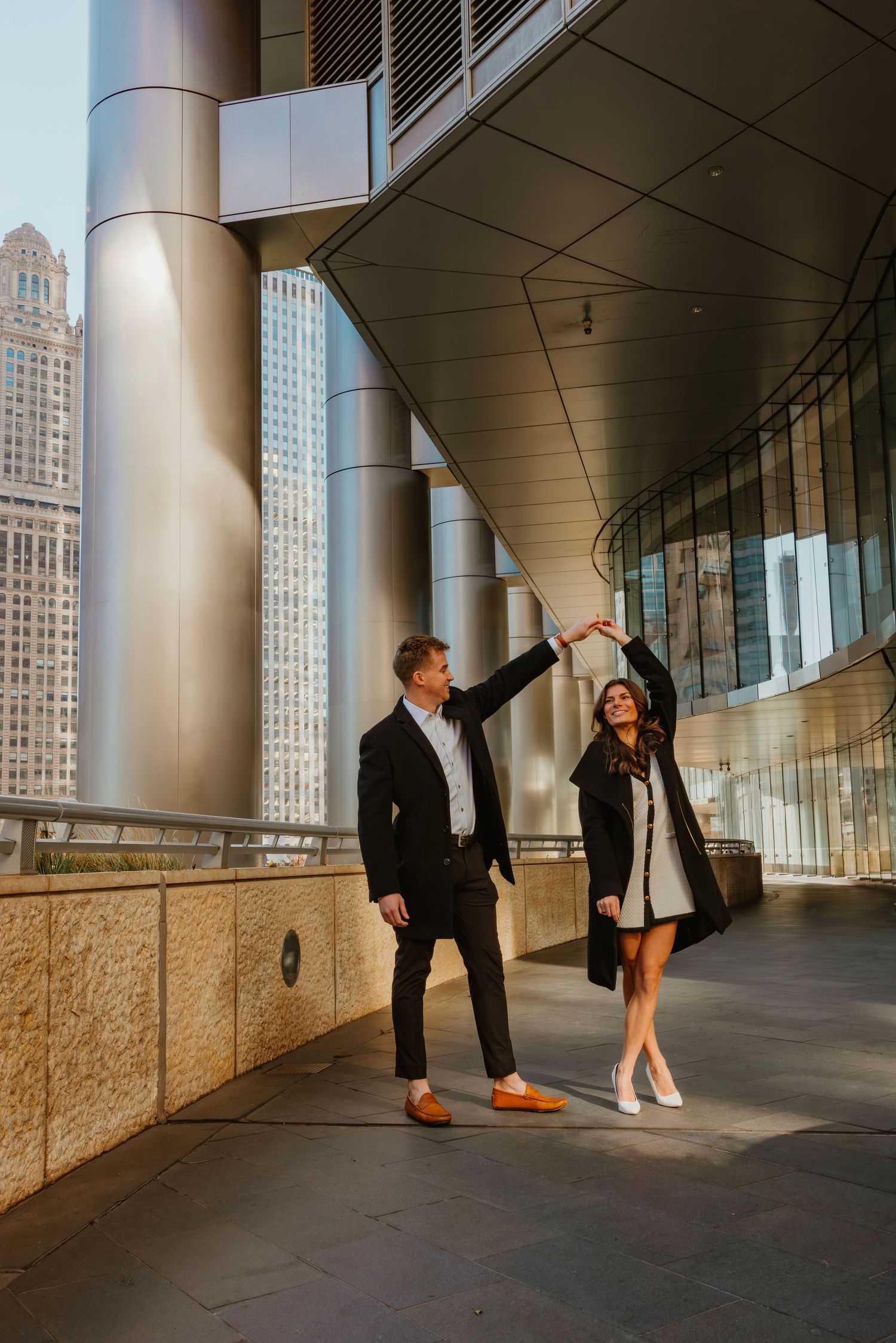 Man spinning woman in white dress under modern building colonnade in Chicago. Outdoor Chicago engagement photos.