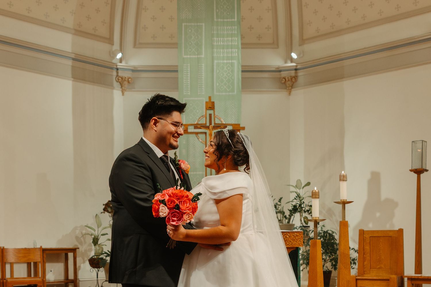 Smiling newlyweds standing at church altar, bride holding vibrant red rose bouquet after wedding ceremony. Chicagoland wedding venues.