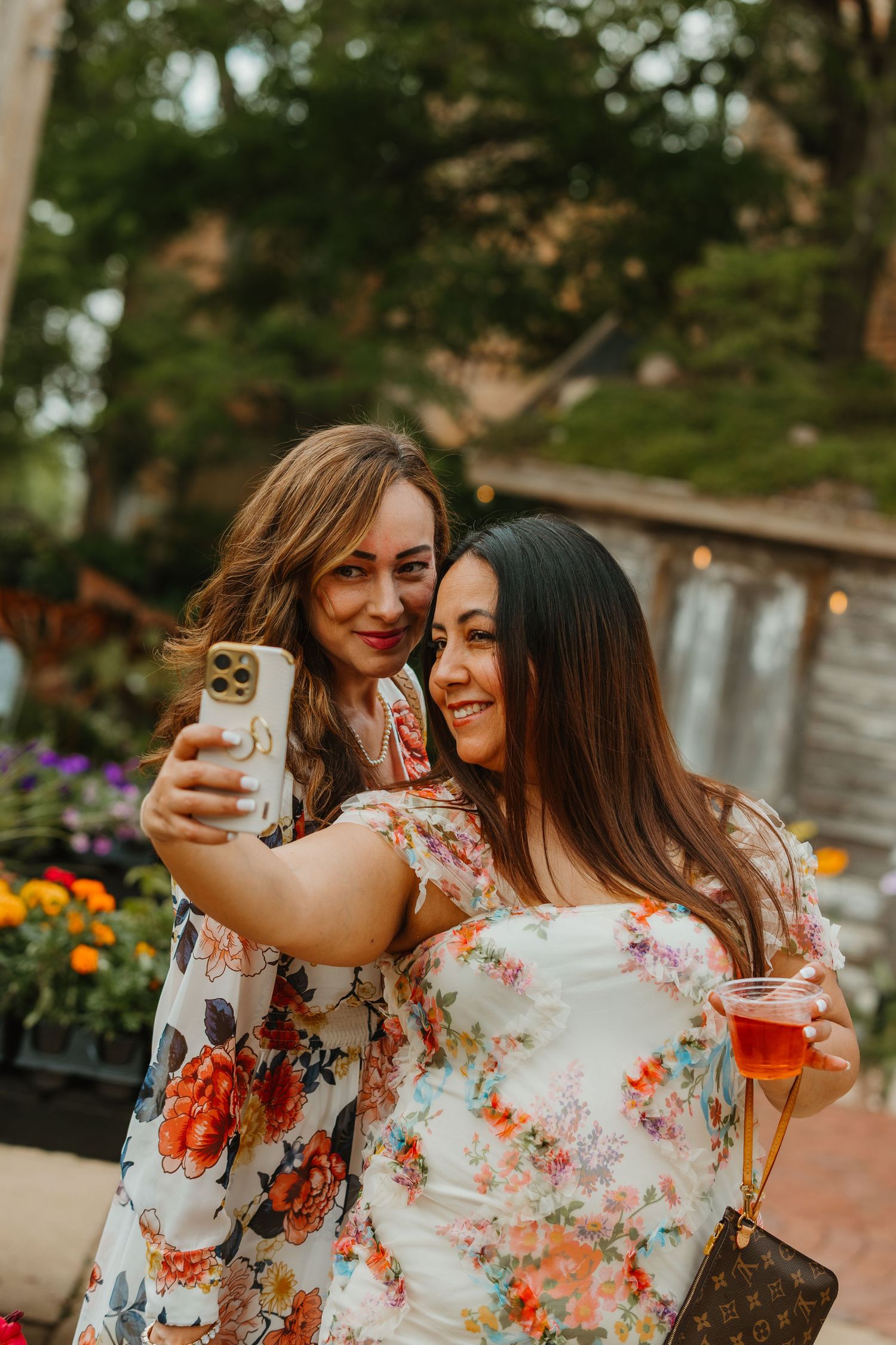 Two women in floral dresses take a selfie outdoors at a garden party, one holding a drink. Chicagoland wedding venues.