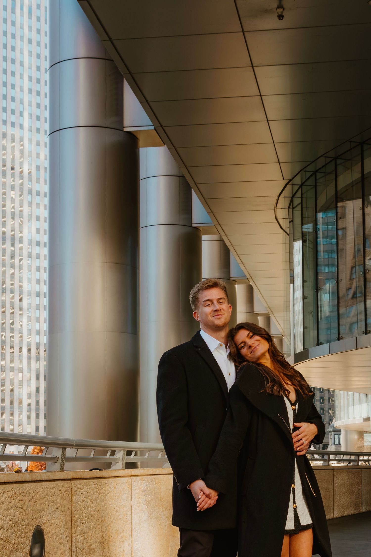 Couple embracing near silver columns with a city skyscraper visible in the background. Outdoor Chicago engagement photos.