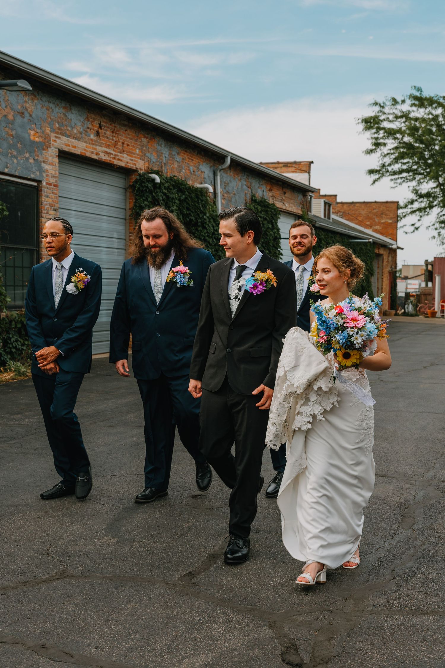 Bride with colorful bouquet walks alongside groom and groomsmen in dark suits on urban street. Chicagoland wedding venues.