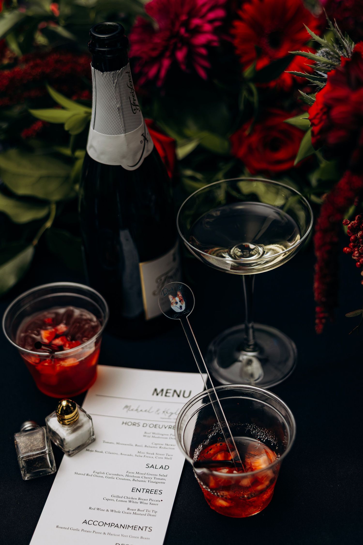 Elegant wedding reception table with champagne bottle, martini glass, red cocktails, a menu card, and lush red floral arrangements.
