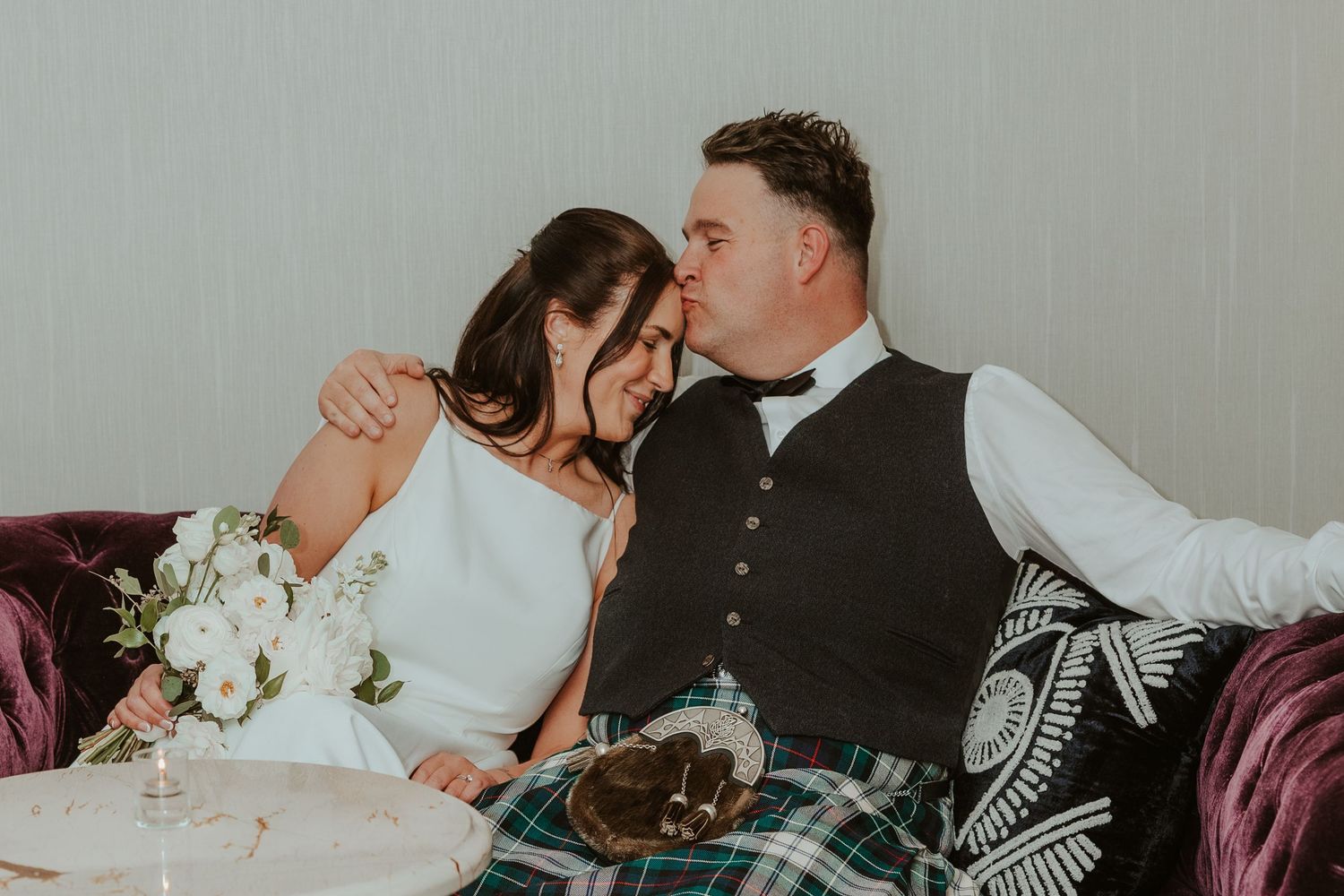 Newlyweds sitting closely on a purple velvet sofa, groom kissing bride's cheek at their wedding reception. Chicago Thompson Hotel wedding.