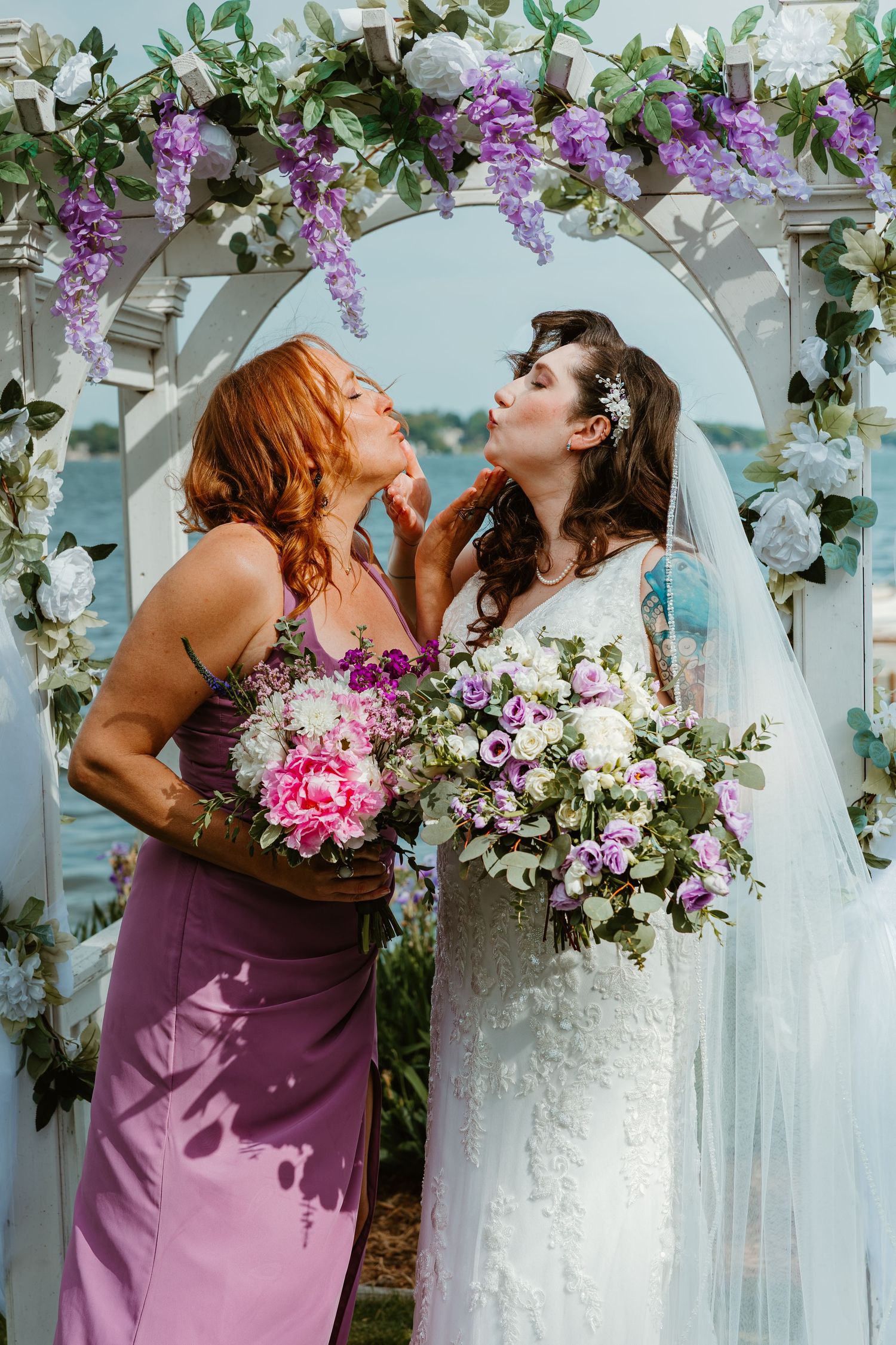 Bride and redhead bridesmaid in lavender dress pose playfully under purple floral wedding arch.