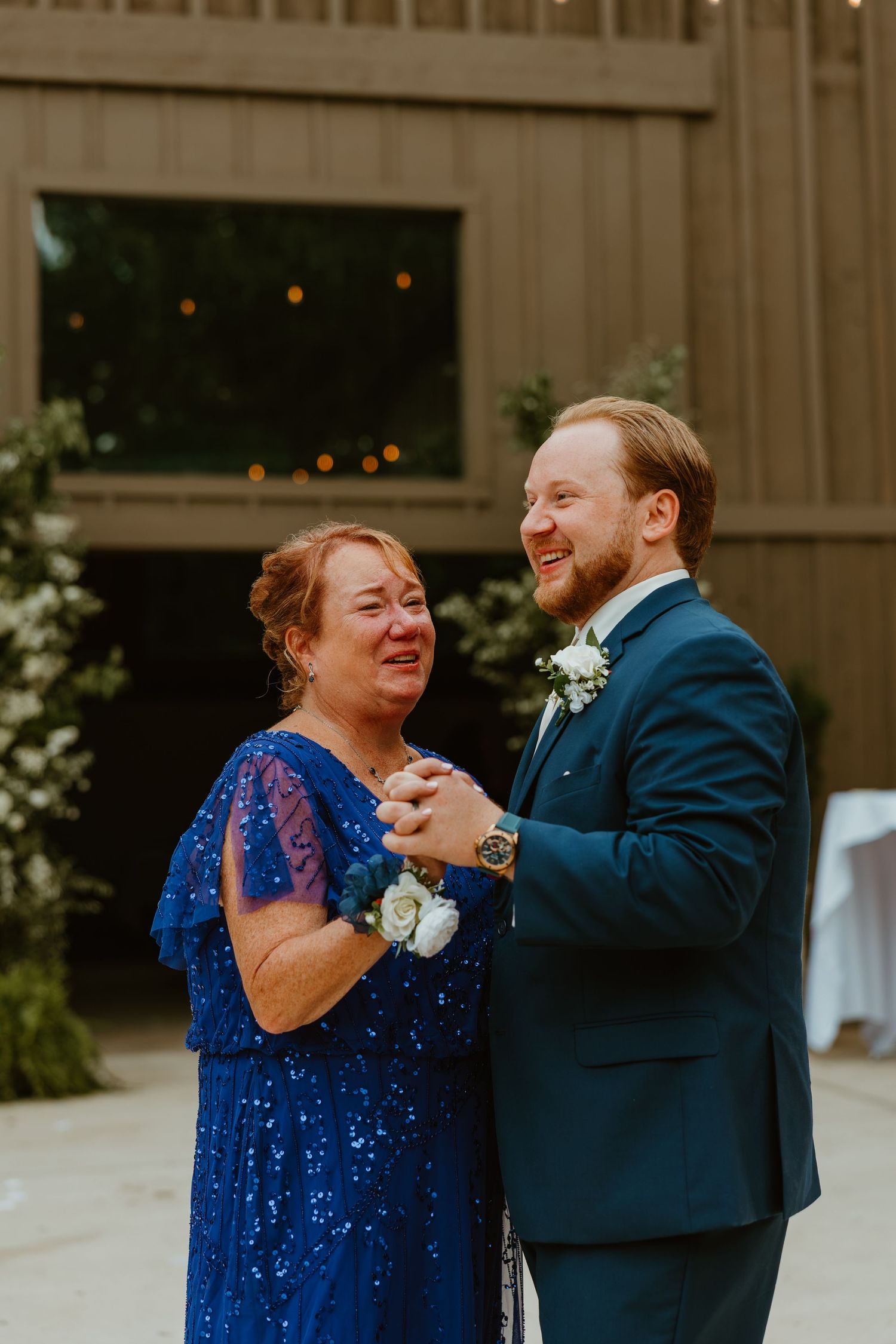 Groom in navy suit laughs joyfully while sharing mother-son dance at rustic barn wedding reception. Northwest Indiana wedding venues.