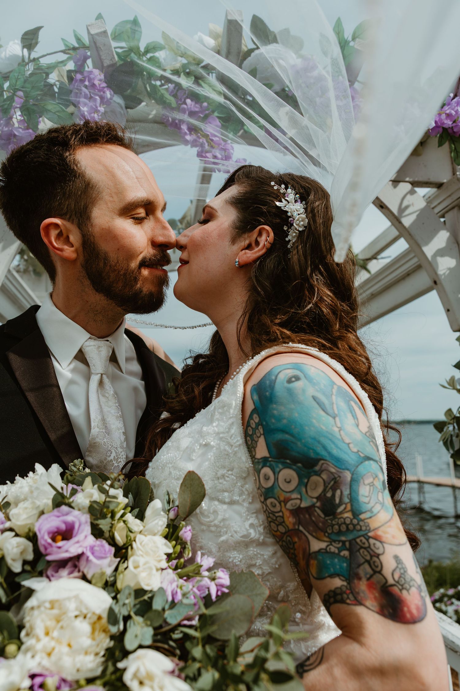 Tattooed bride and bearded groom share first kiss under floral arch with purple wisteria at lakeside wedding.