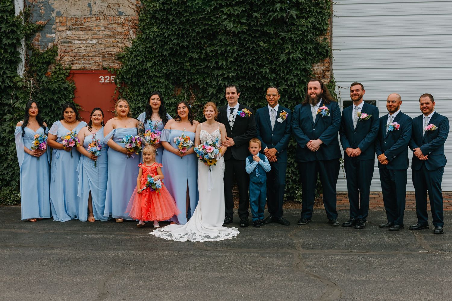 Large wedding party with bridesmaids in blue, groomsmen in navy, and young ring bearer poses formally outdoors. Chicagoland wedding venues.