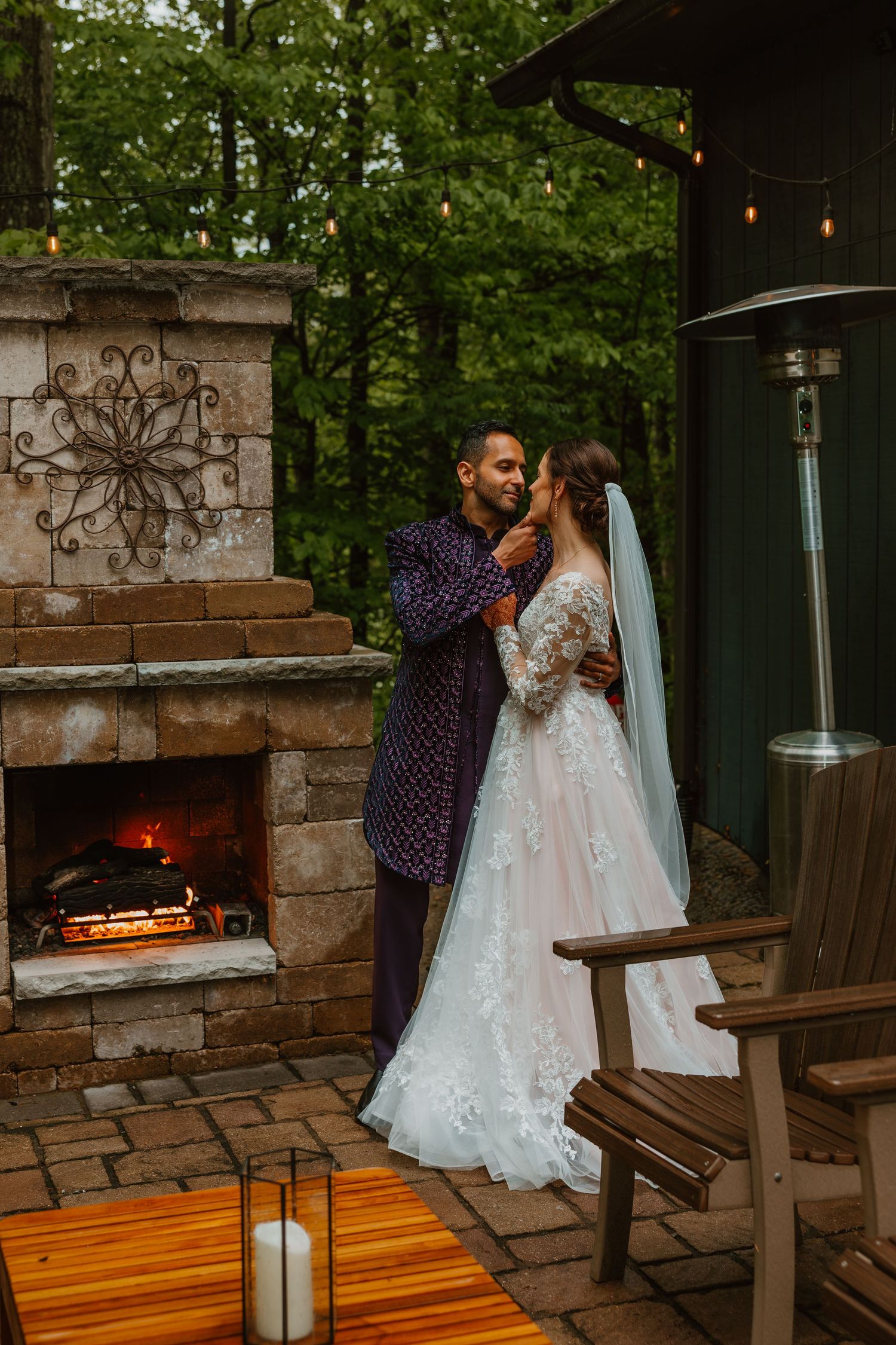 Bride and groom sharing an intimate moment by a glowing stone fireplace on a rustic patio. Documentary style wedding photos.