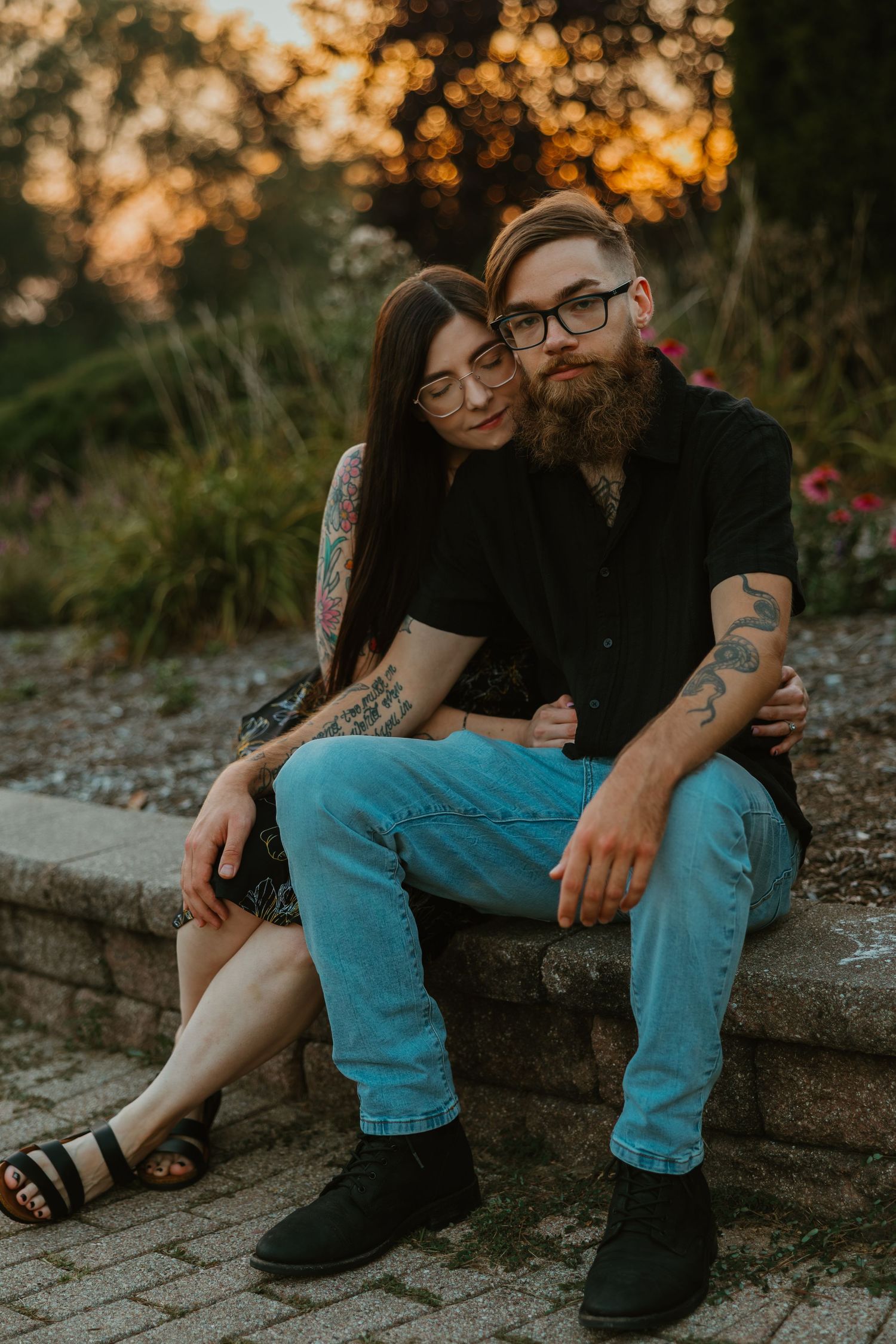Couple with tattoos sitting on stone steps at sunset, woman leaning on bearded man's shoulder in a garden. Chicagoland engagement photos.