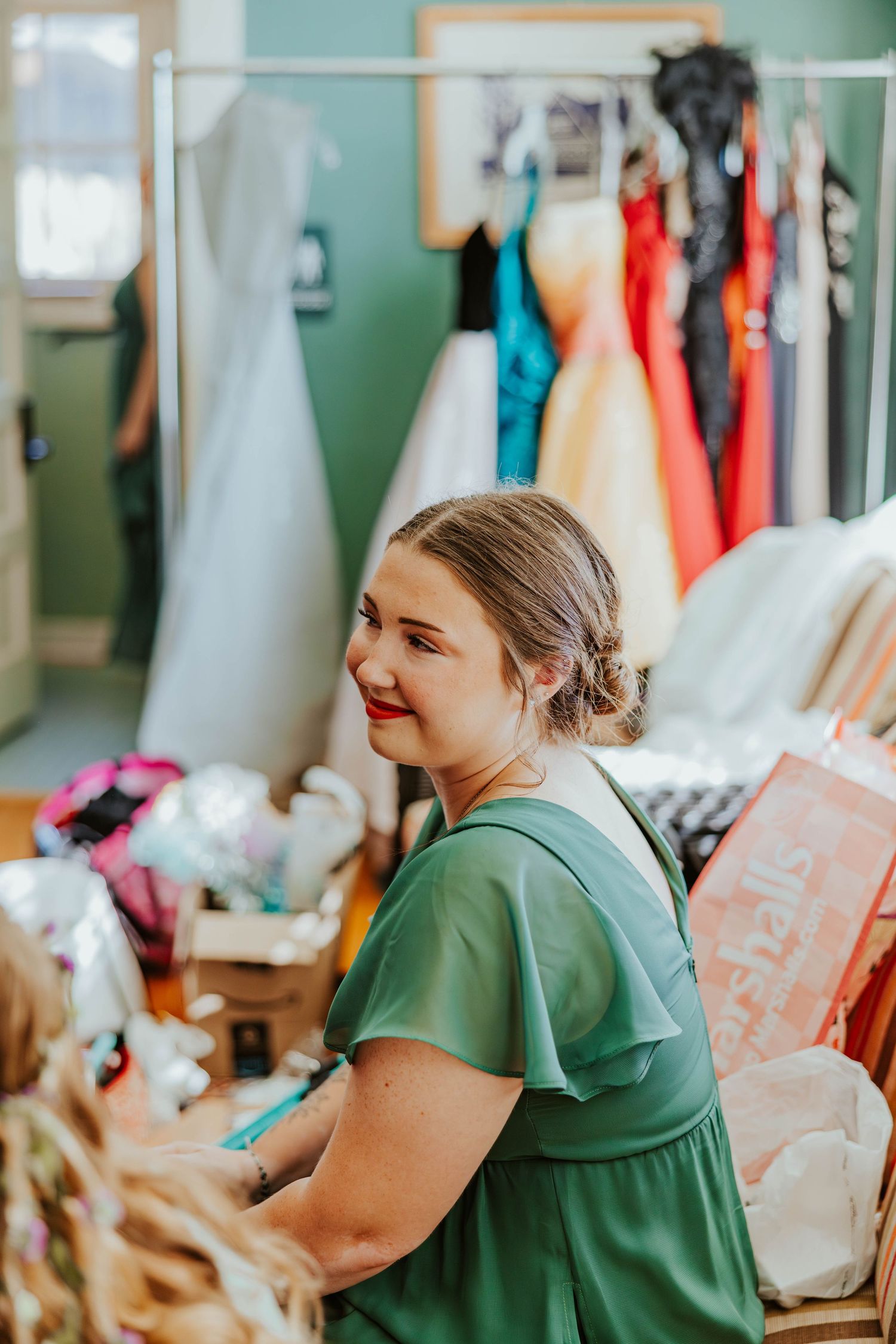 Smiling bridesmaid in green dress stands near clothing rack filled with colorful wedding party gowns.