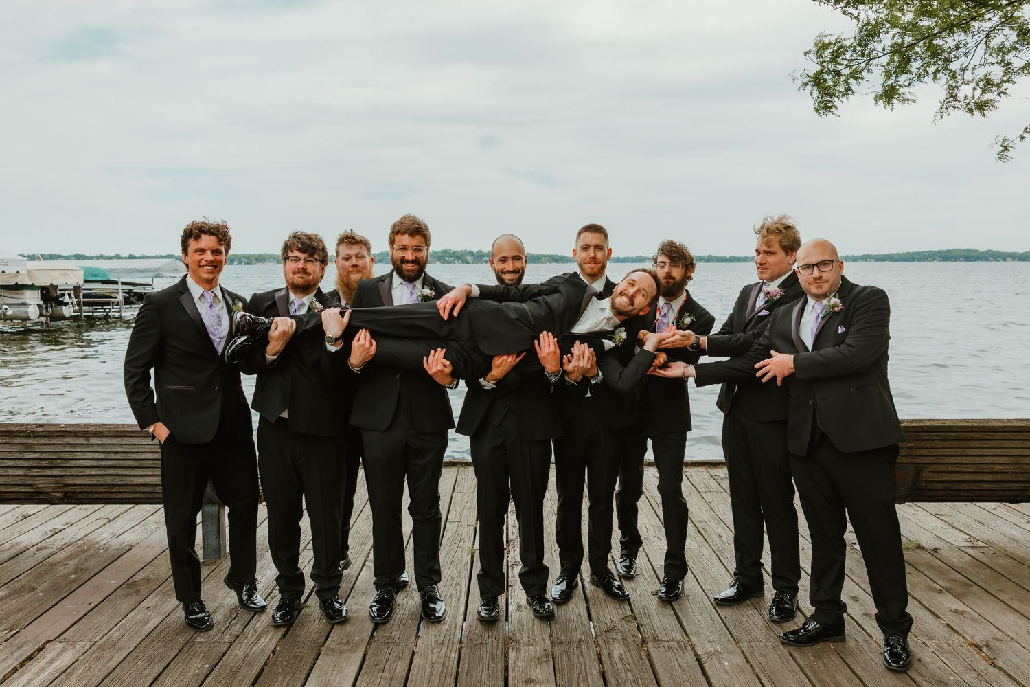 Groomsmen in black suits posing playfully on a wooden dock with a lake in the background.