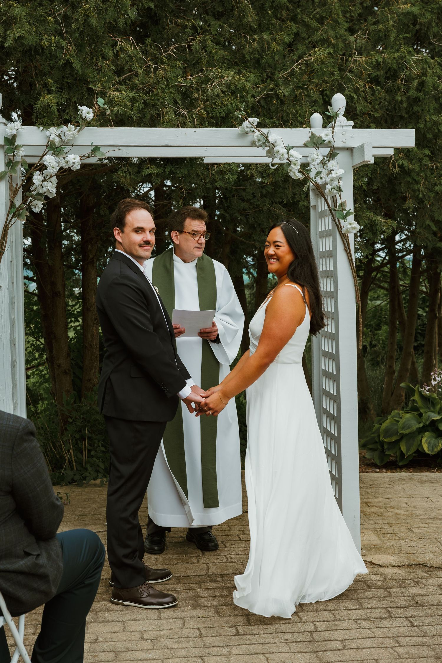 Bride and groom hold hands during outdoor wedding ceremony under white floral arch with officiant. Michigan elopement.