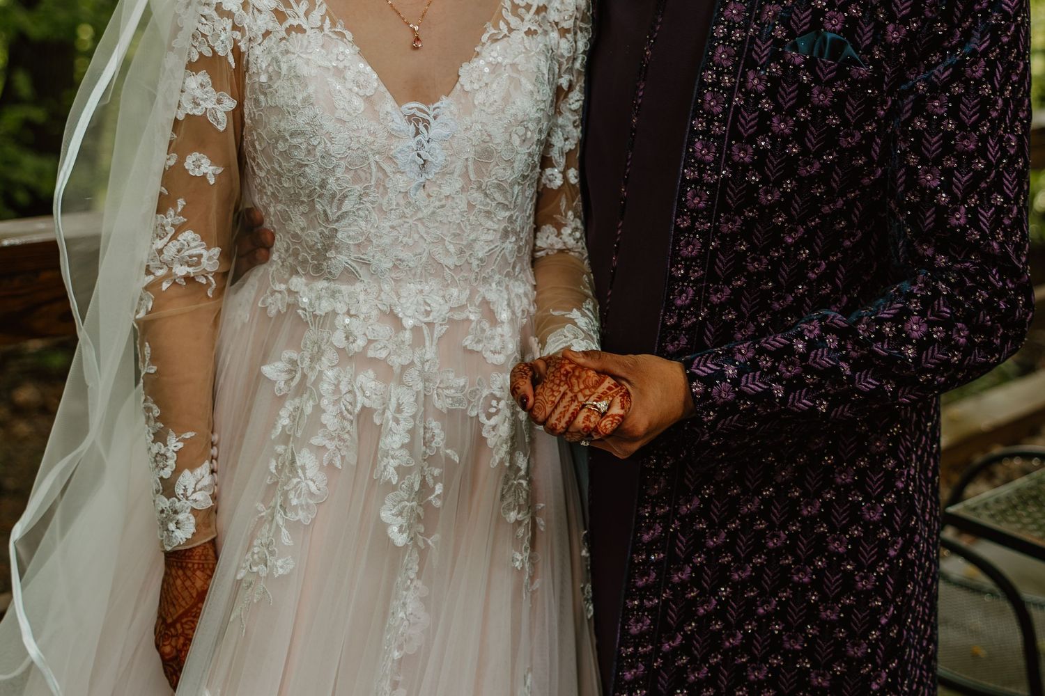 Close-up of bride in lace gown and groom in floral suit holding hands with henna-adorned hands. Documentary style wedding photos.