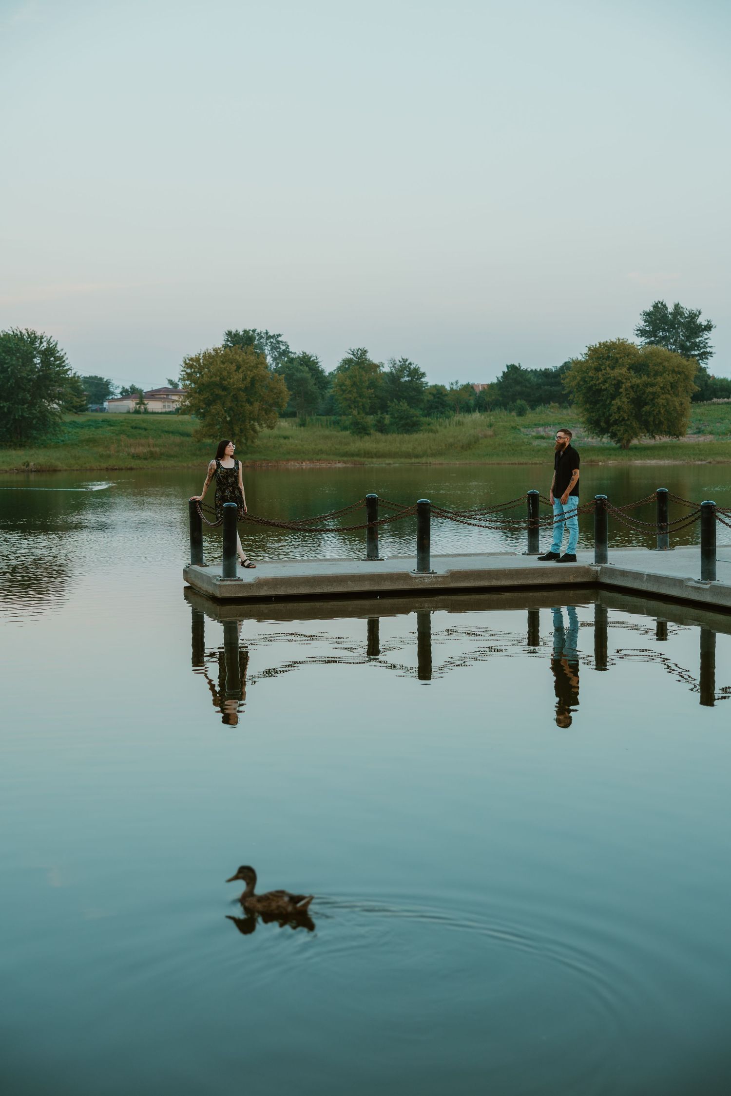 Two people standing apart on a lakeside dock at twilight, with a duck swimming in the reflection below. Chicagoland engagement photos.