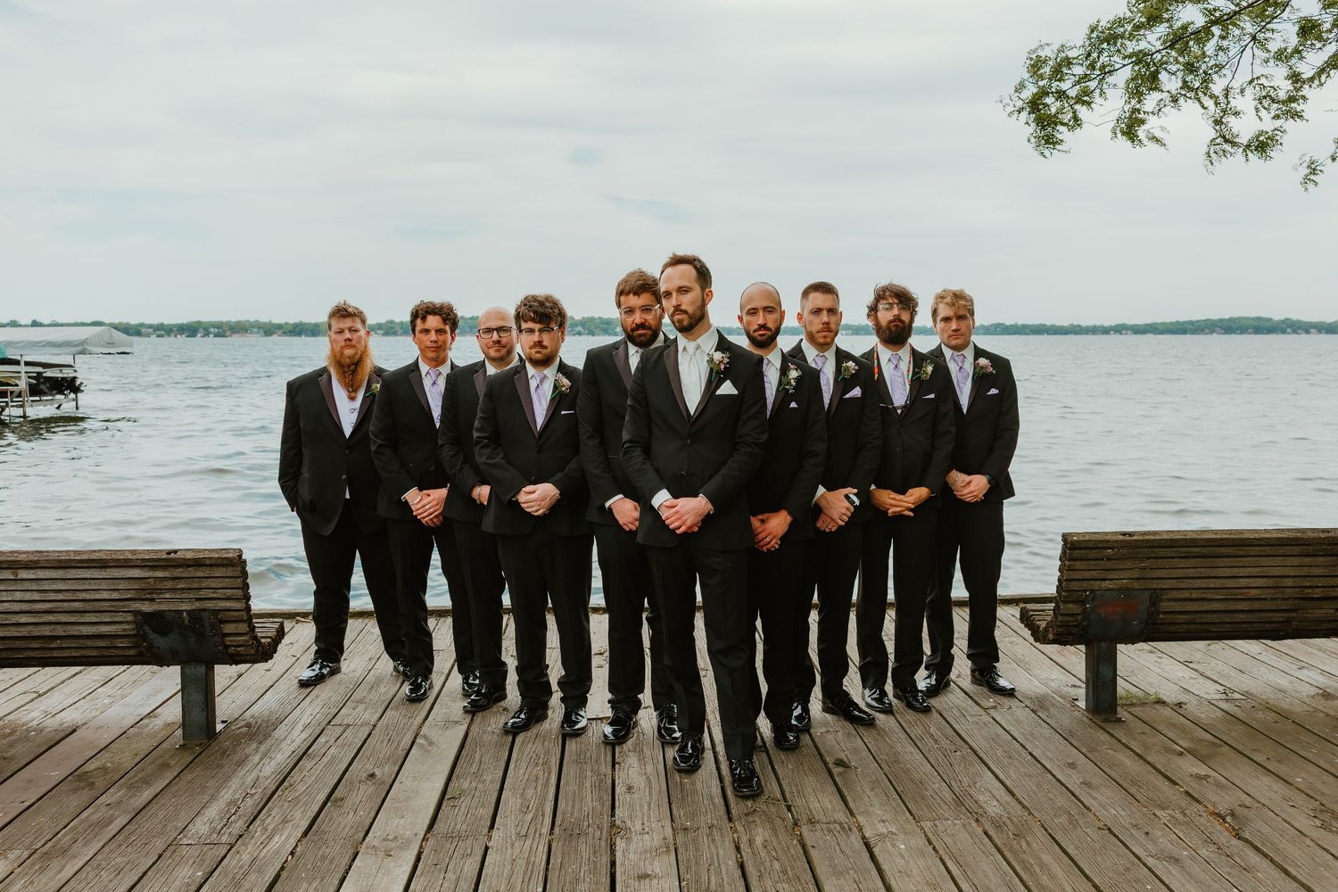 Groom with groomsmen in black suits standing formally on a lakeside wooden dock before the wedding.