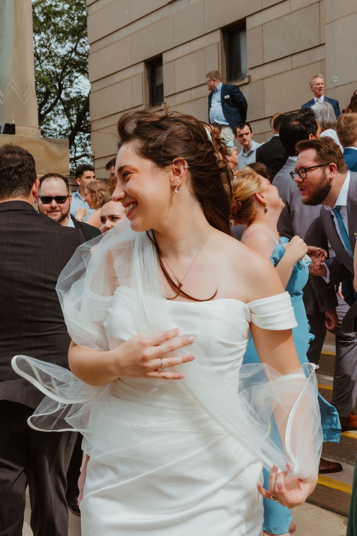 Smiling bride in white off-shoulder wedding gown holding veil surrounded by wedding guests outdoors.
