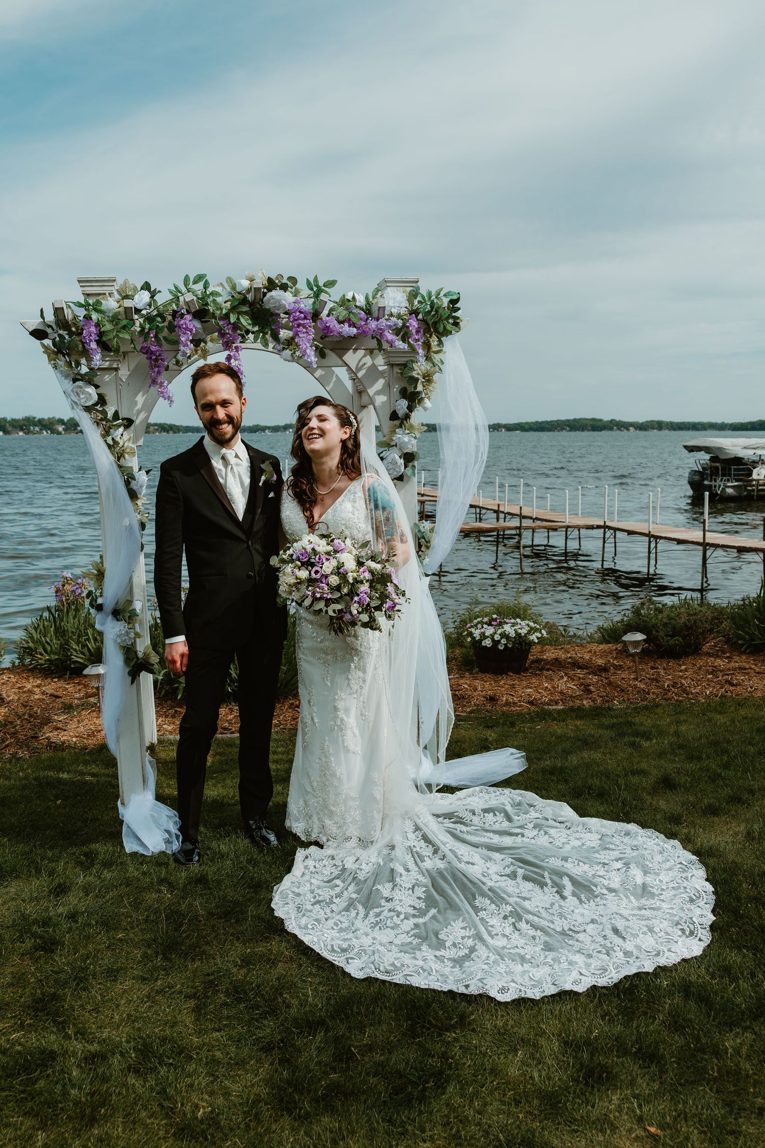 Smiling bride in lace gown and groom stand under purple floral arch with lake and dock behind them.