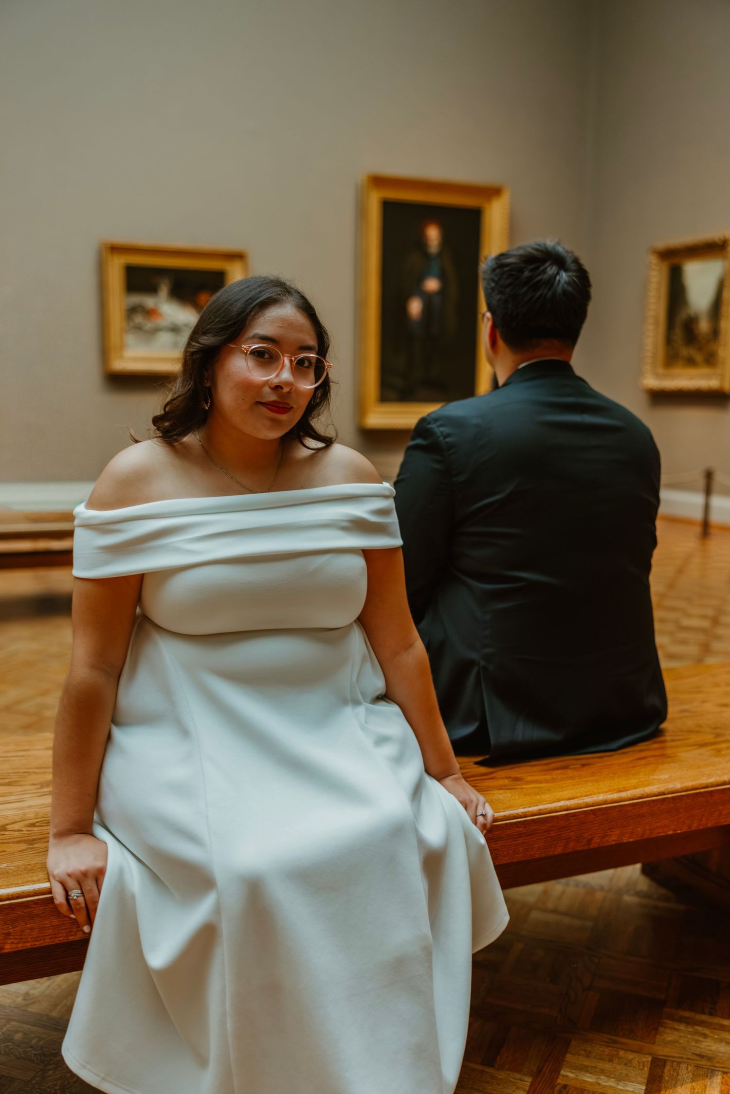 Bride in off-shoulder white dress sits on museum bench smiling, groom in black suit views artwork behind her. Art Institute engagement photo