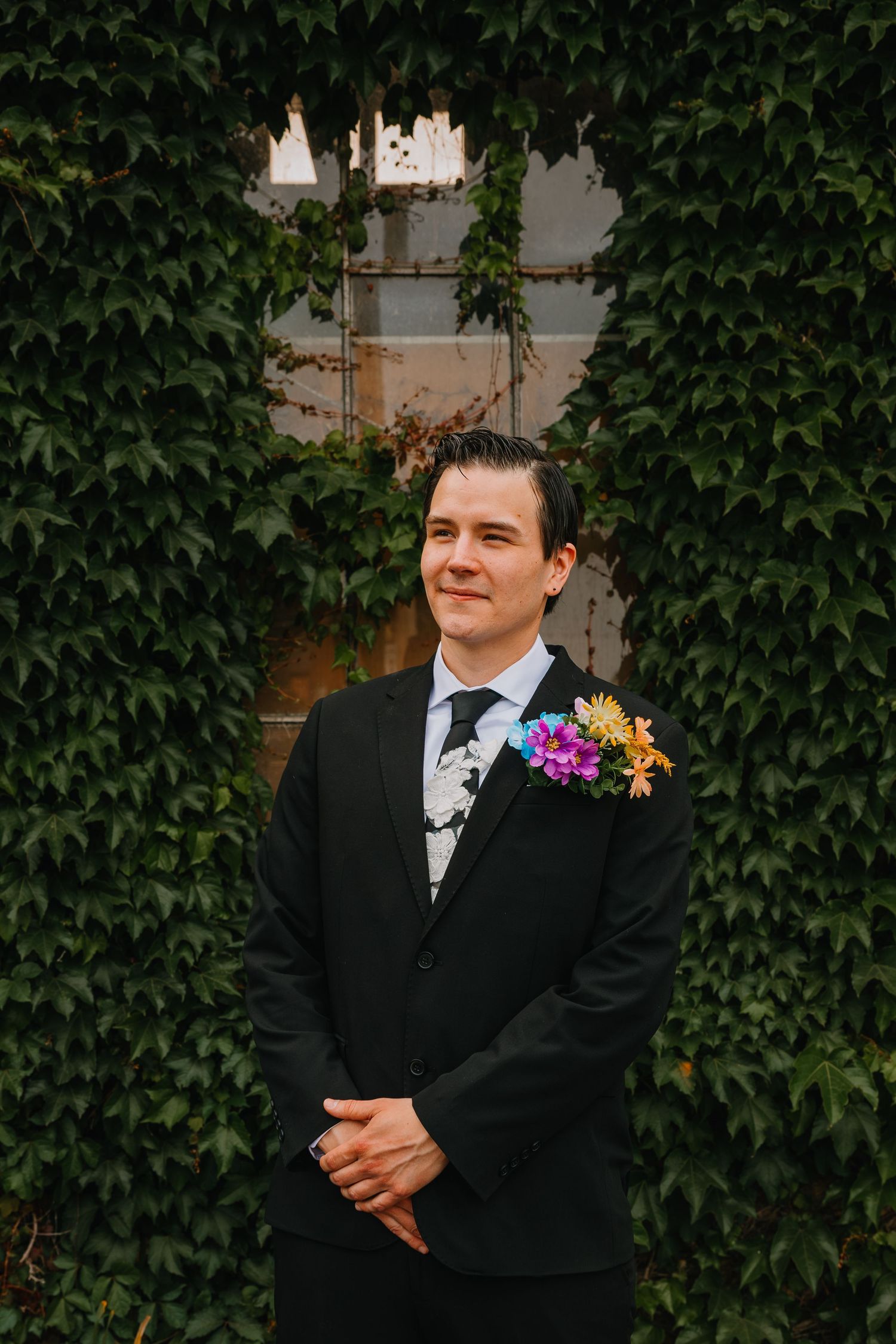 Groom in black suit with colorful boutonniere stands confidently before lush ivy-covered wall outdoors. Chicagoland wedding venues.