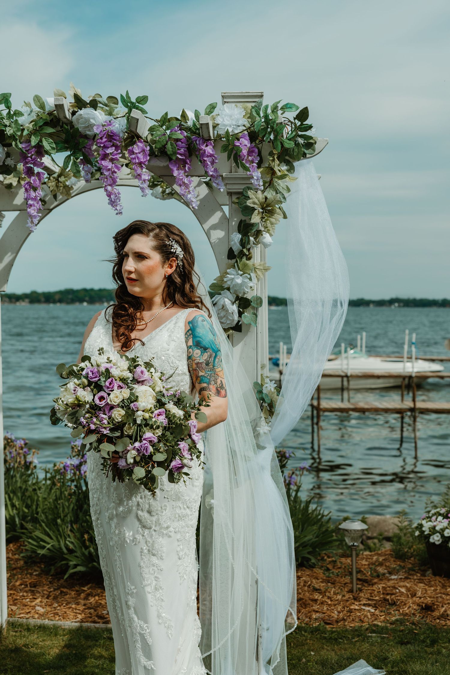Bride in fitted lace wedding dress holds purple and white bouquet under a floral arch lakeside.