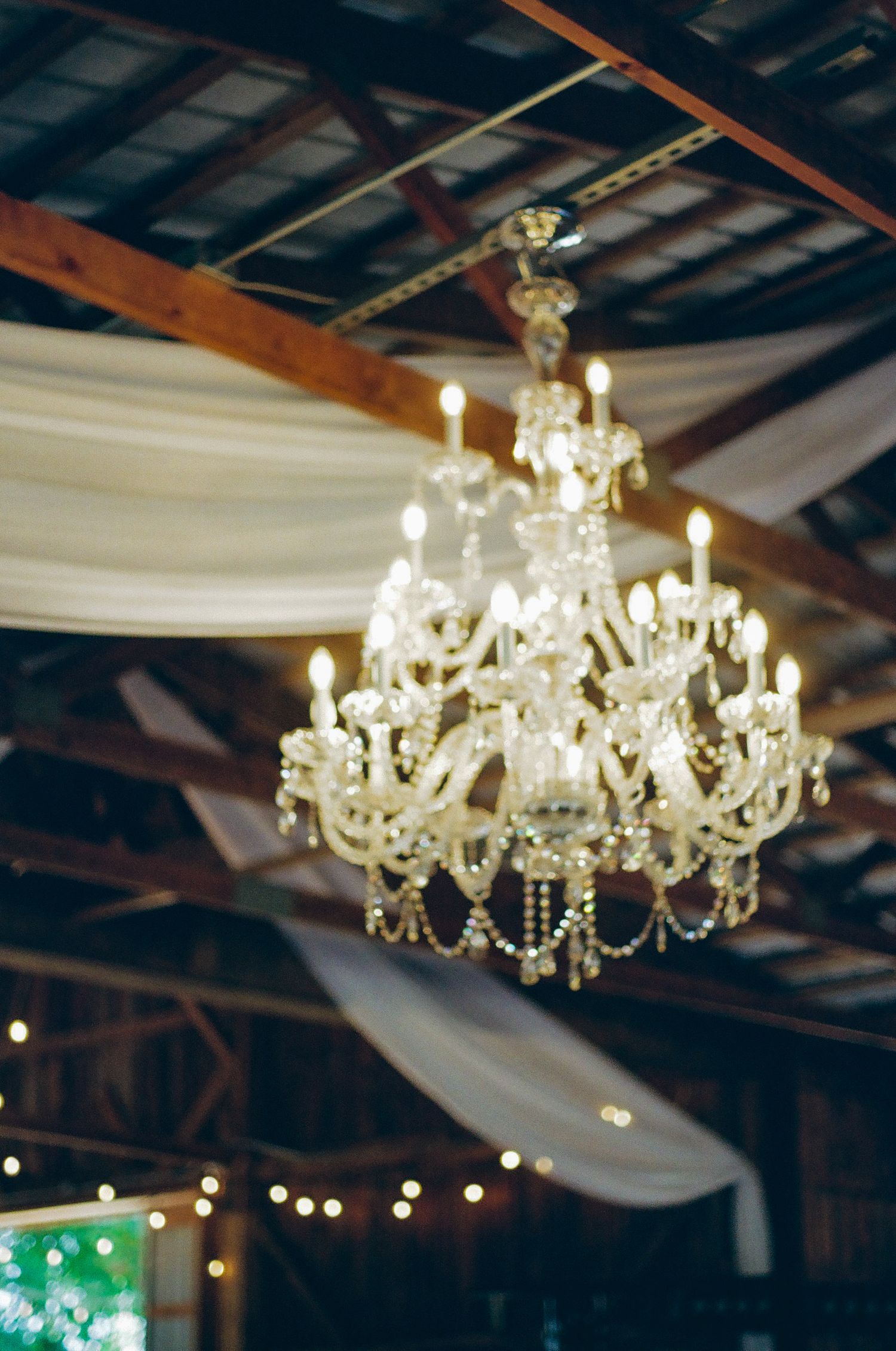 Elegant crystal chandelier with candle lights hanging from rustic wooden ceiling with white draped fabric at wedding venue.