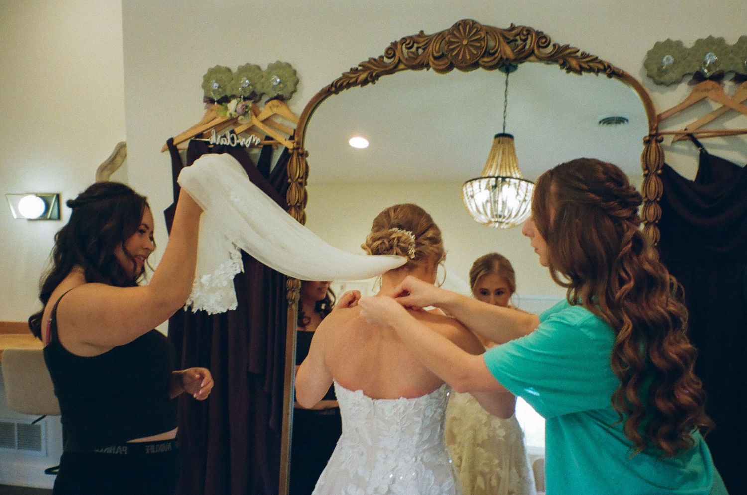 Bridesmaids helping bride into lace wedding dress in front of ornate gold mirror in bridal suite.