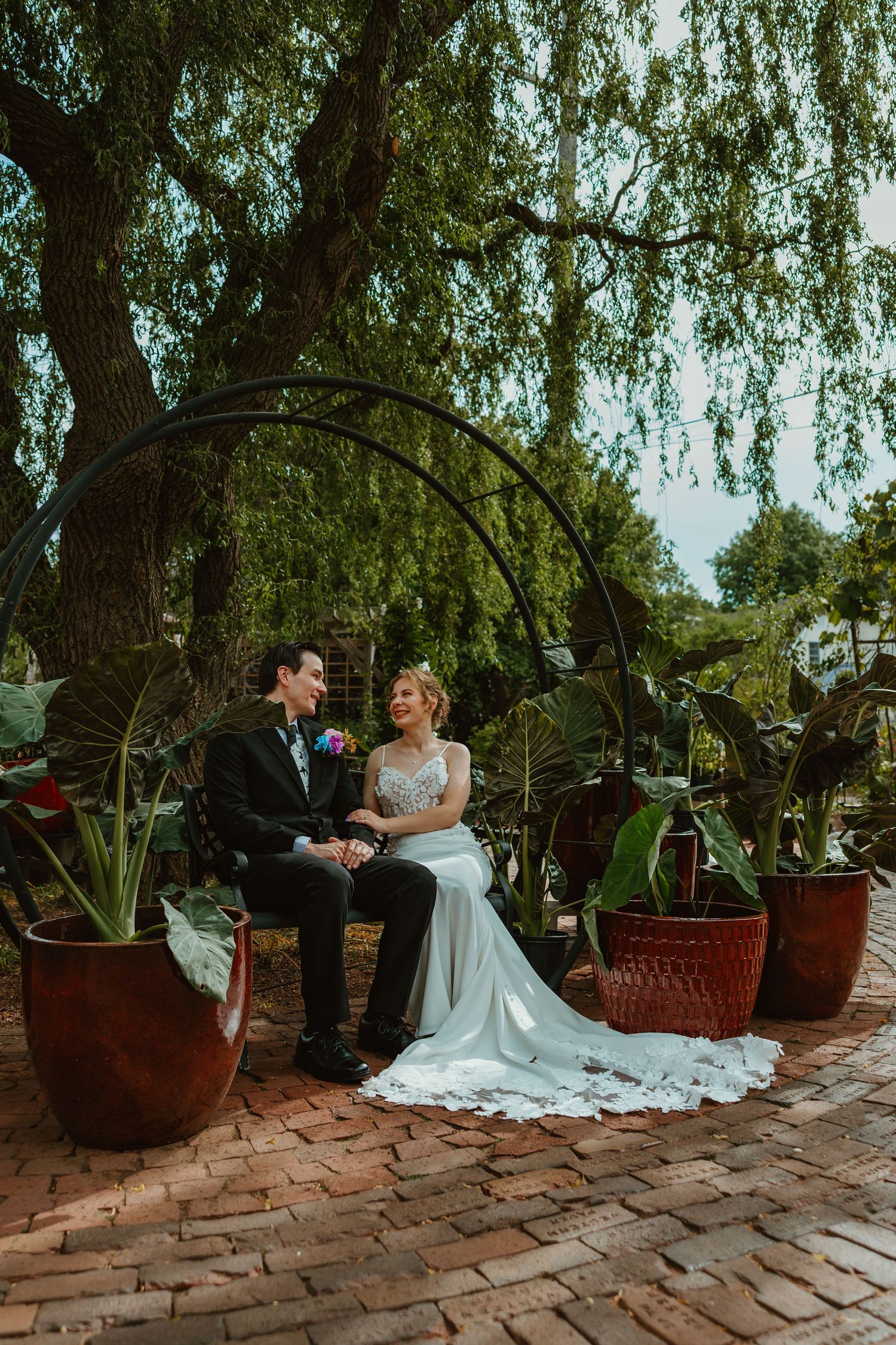 Wedding couple sitting on a bench under a willow tree arch, smiling at each other in a garden setting. Chicagoland wedding venues.
